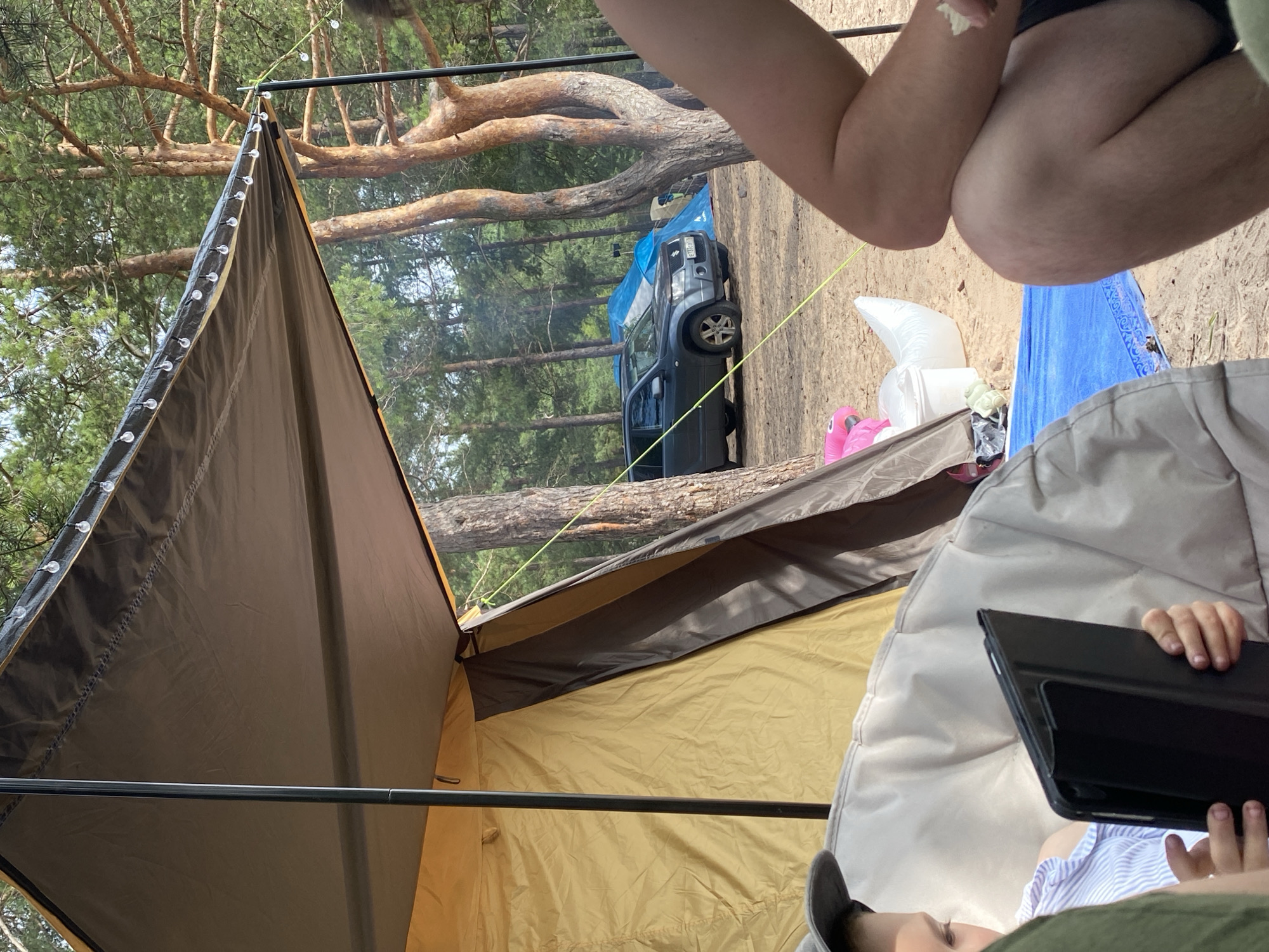 The image depicts a campsite in Baltiiskoe, Russia, with a focus on a partially set-up tent and awning. A dark grey SUV is parked behind the tent. Two blue tents are visible in the background, along with several pine trees. In the foreground, a child wearing a grey cap and a striped shirt is seated on a large, light-colored beanbag chair, holding a black tablet. To the right, the arm and leg of an adult are visible, with the adult appearing to hold a piece of food. A blue tarp lies on the sandy ground near the beanbag chair, and a white bucket and a deflated pink inflatable object are also present. The scene suggests a camping trip in a wooded area.