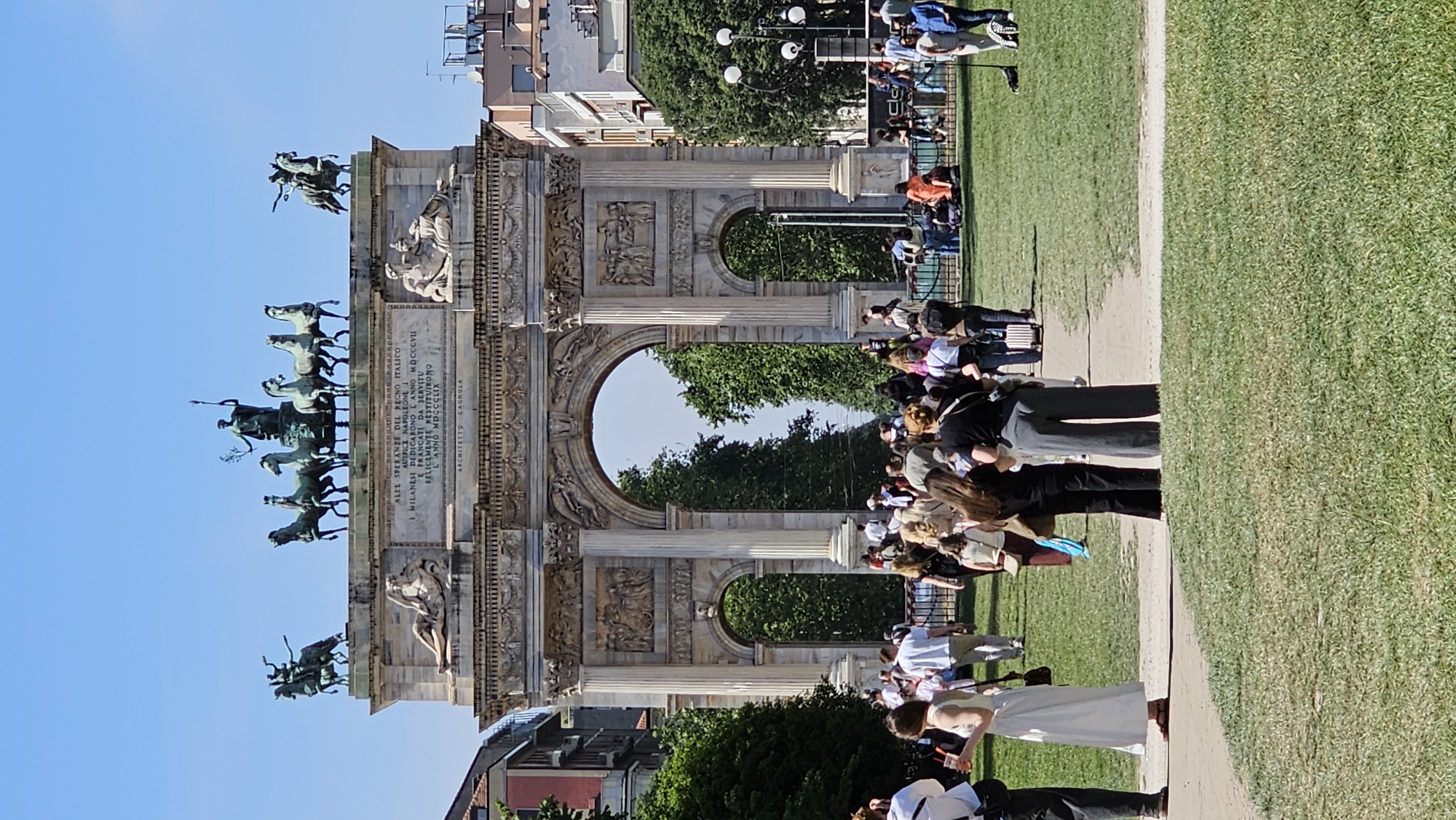 The image shows the Arco della Pace (Arch of Peace) in Milan, Italy. The neoclassical triumphal arch, topped with a bronze sculpture of horses and figures, dominates the scene. People are walking and standing around the arch, including a woman in a white dress and a man in a black suit, potentially taking wedding photos. A grassy area borders a pedestrian path. The sky is blue. Buildings and trees are visible in the background.