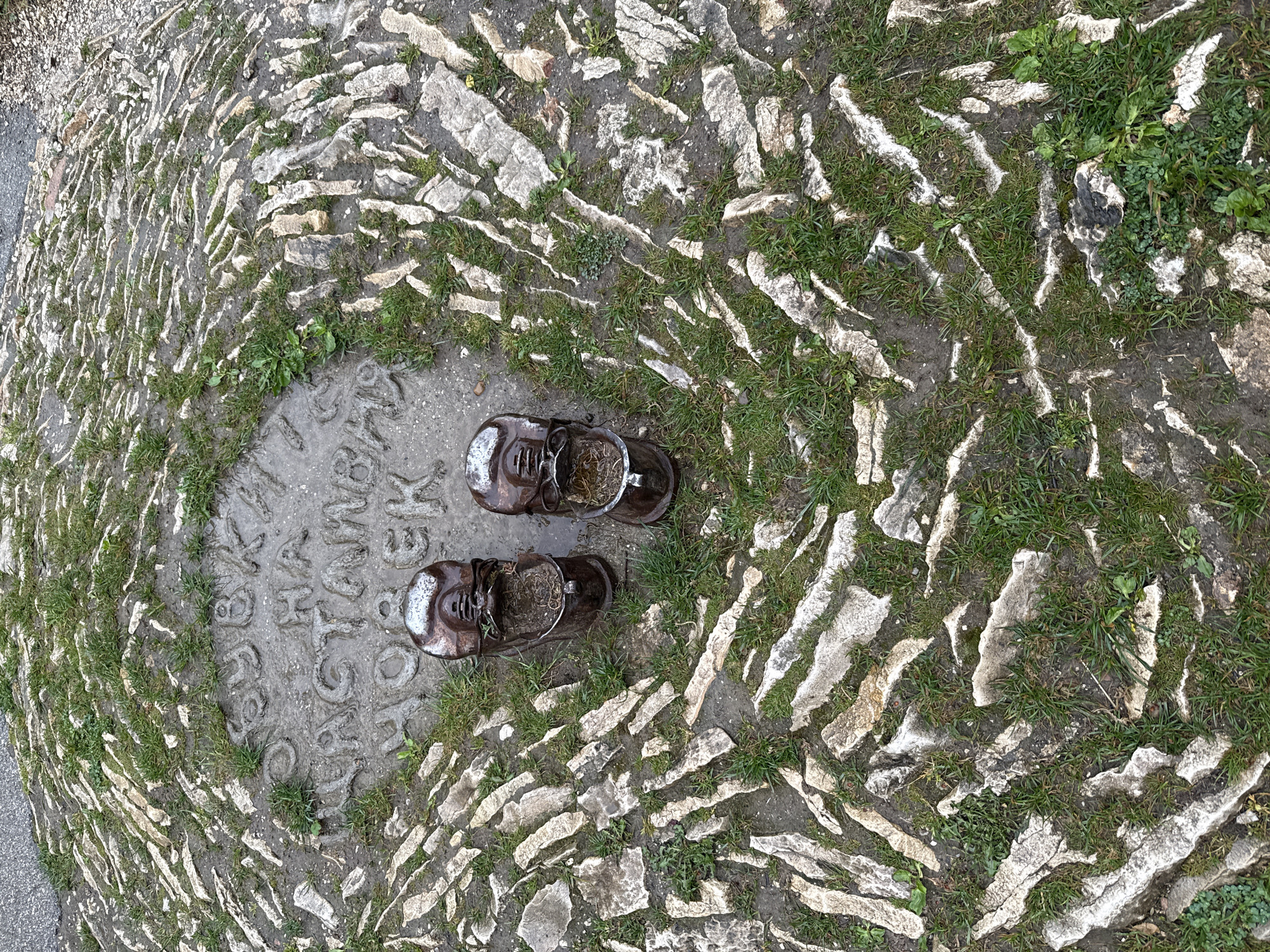 A top-down view shows a circular stone surface with inset text. Two bronze-colored, antique-style shoes filled with straw are placed centrally on the stone, facing in opposite directions. The circular area is constructed from flat, irregular stone pieces with grass growing between them. The location is near Lukovit, Bulgaria, as indicated by the text "CT. ЛУКОВИТЪ БЪЛГАРИЯ", which is partially visible on the central stone. The environment appears to be outdoors, possibly a monument or landmark in a natural setting.