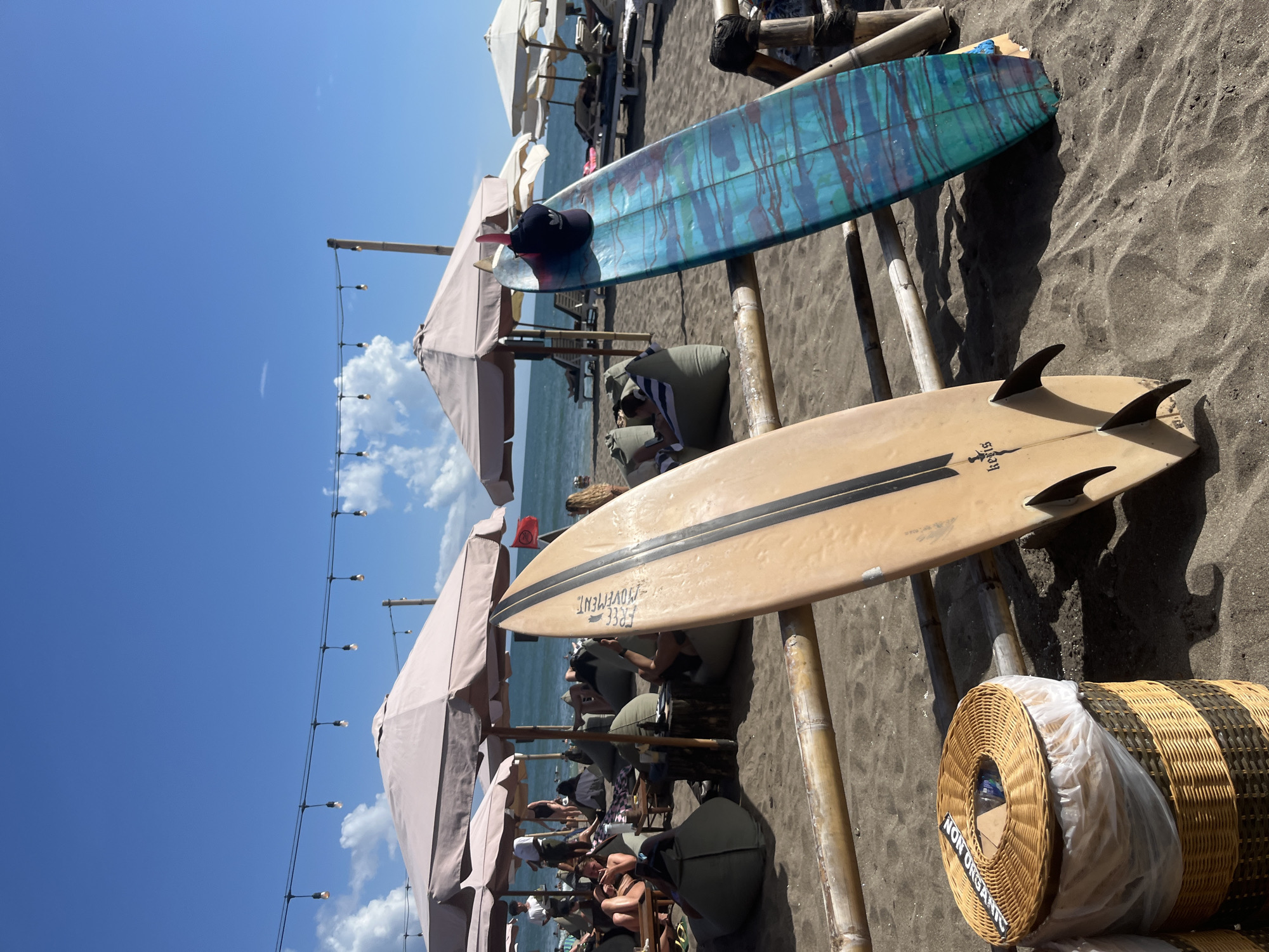 The image depicts a beach scene in North Kuta, Indonesia. Two surfboards are laid horizontally across bamboo supports. The top surfboard has a blue and green marbled pattern, with a black hat resting on it. The bottom surfboard is tan with black stripes and three fins. Underneath the surfboards, people are seated on chairs and beanbag-like cushions under parasols. A coiled roll of woven material, labeled "NON ORGANIC", sits at the lower right of the frame. A string of lights is visible on the left, extending towards a blue sky with scattered clouds. The beach is covered in dark-colored sand. The ocean is visible in the background.