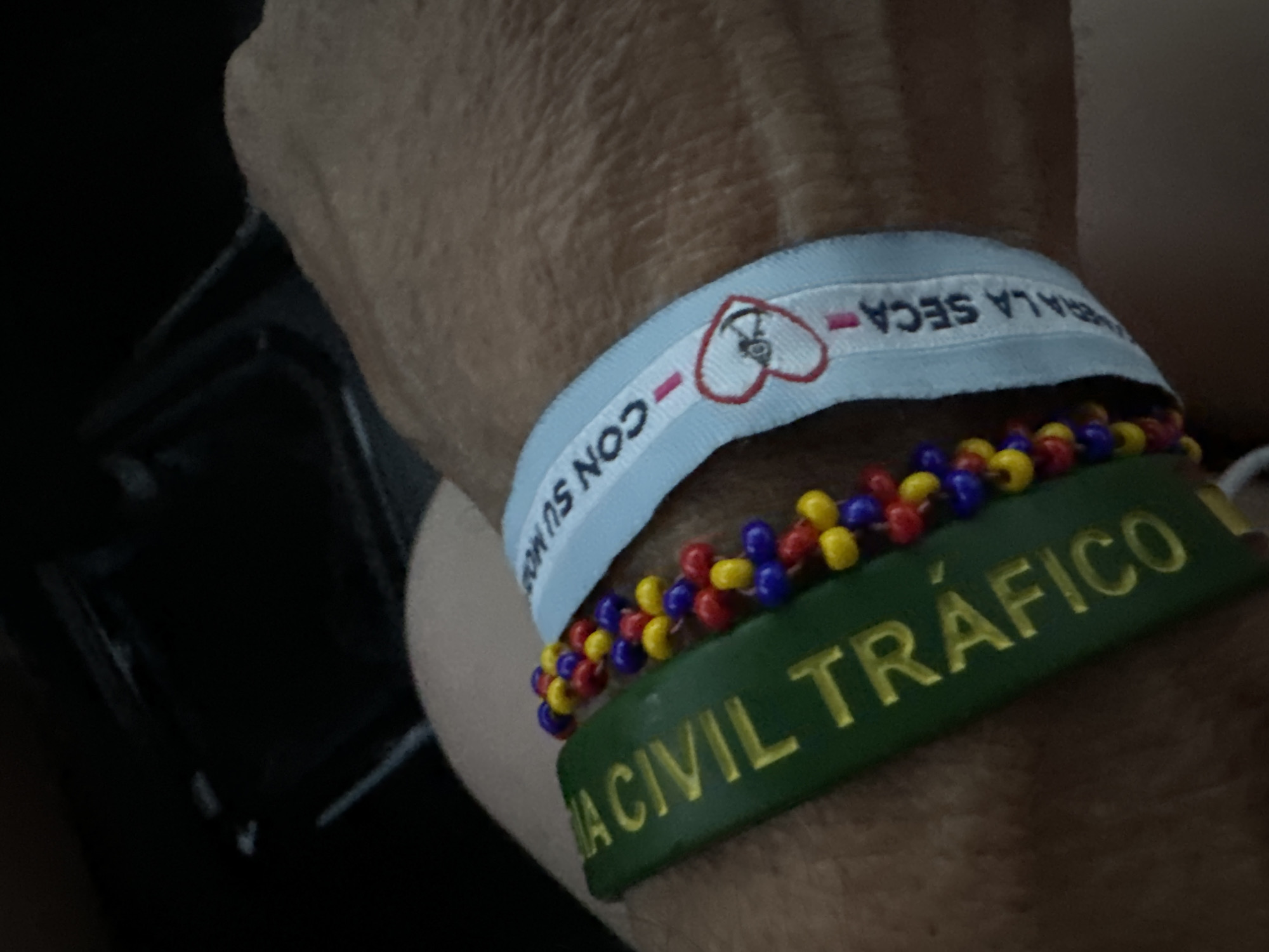 The image shows a close-up of a person's wrist wearing two bracelets. One is a green fabric band with yellow text that reads "A CIVIL TRAFICO". The other is a blue fabric band with white text and a red heart logo containing an anchor. The text on the blue band reads "CON SU MANO... LA SECA". A beaded bracelet with red, yellow, and blue beads is visible between the two fabric bands. The background is dark and out of focus, suggesting an indoor setting. The scene is a close-up of jewelry on a wrist, with no discernible city or environmental details.