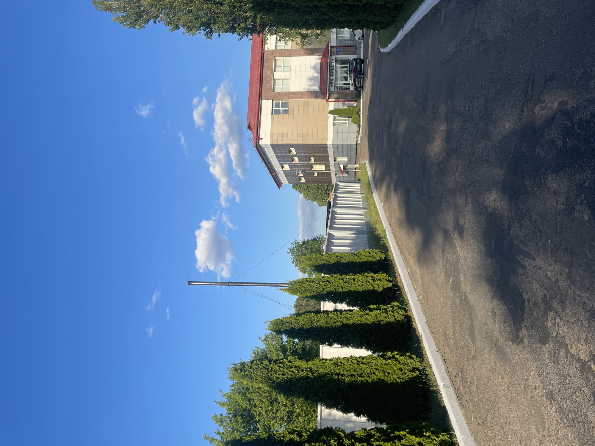 Exterior view of the "VK Brest" building in Brest, Belarus, next to an asphalt driveway with bordering shrubbery, under a blue sky with scattered clouds. No people are visible. A flag pole stands near the shrubs. A black car is parked in front of the building's entrance. The building has a red roof and multi-toned facade.