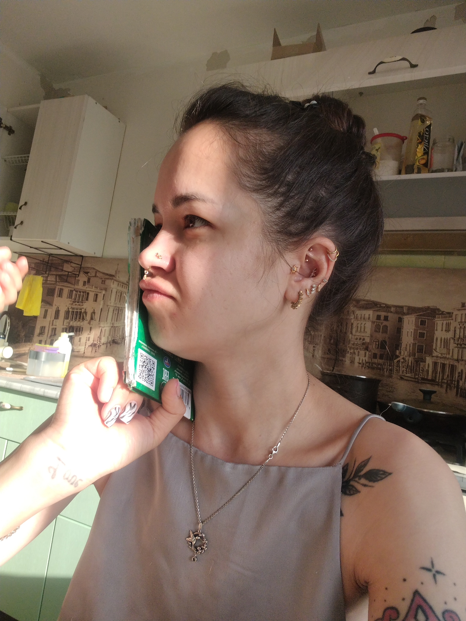 A medium shot captures a woman in a brightly lit kitchen. She is holding a green, rectangular object to her ear, seemingly mimicking a phone call. She has multiple piercings on her ear and nose. Her dark hair is pulled back in a bun. She wears a light-colored, sleeveless top and a necklace. Tattoos are visible on her arm and shoulder.
The kitchen features light green cabinets and a backsplash with a Venetian-style cityscape. A white cabinet is visible in the upper left. A shelf above the backsplash holds various items including a bottle of cooking oil. The location is Noviy, Russia.