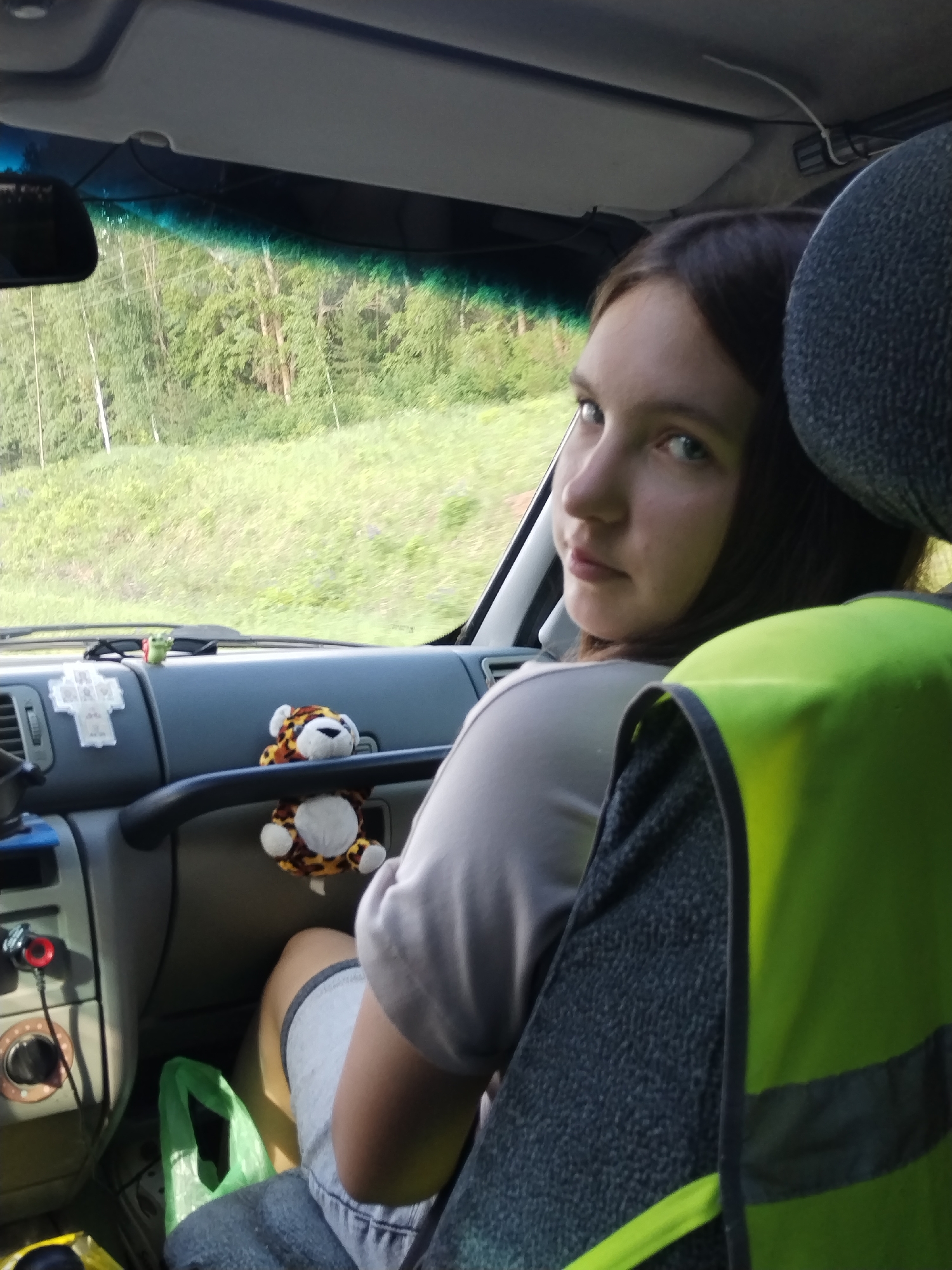 A medium shot captures a young female passenger seated in the front of a car, turning her head and torso to look back over her right shoulder directly at the camera. She has long, dark hair, light-colored eyes, and a neutral, slightly contemplative expression. She is wearing a light-colored, short-sleeved top and light-toned shorts. A bright lime green/yellow vest is draped over the seat back on the right, partially visible.
The car's interior features a dark grey headrest and a light-grey dashboard. A small plush tiger toy hangs from the passenger-side grab handle on the dashboard. To its left, a white cross-shaped ornament, likely an air freshener or religious icon, is affixed to the dashboard, displaying the Cyrillic text "НИКА" (NIKA). Sunglasses and a small green frog figurine are also visible on the dashboard. On the driver's side, parts of the car's controls, including a circular knob and some wiring, are visible. The windshield has a blue-green tint along its top edge.
Through the front windshield, a bright, verdant landscape is visible, characterized by numerous green trees and lush vegetation, suggesting a rural or forested area. The clear and bright lighting indicates it is daytime, likely late morning or afternoon. The image captures a moment during a car journey in the area of Фоки, Russia.