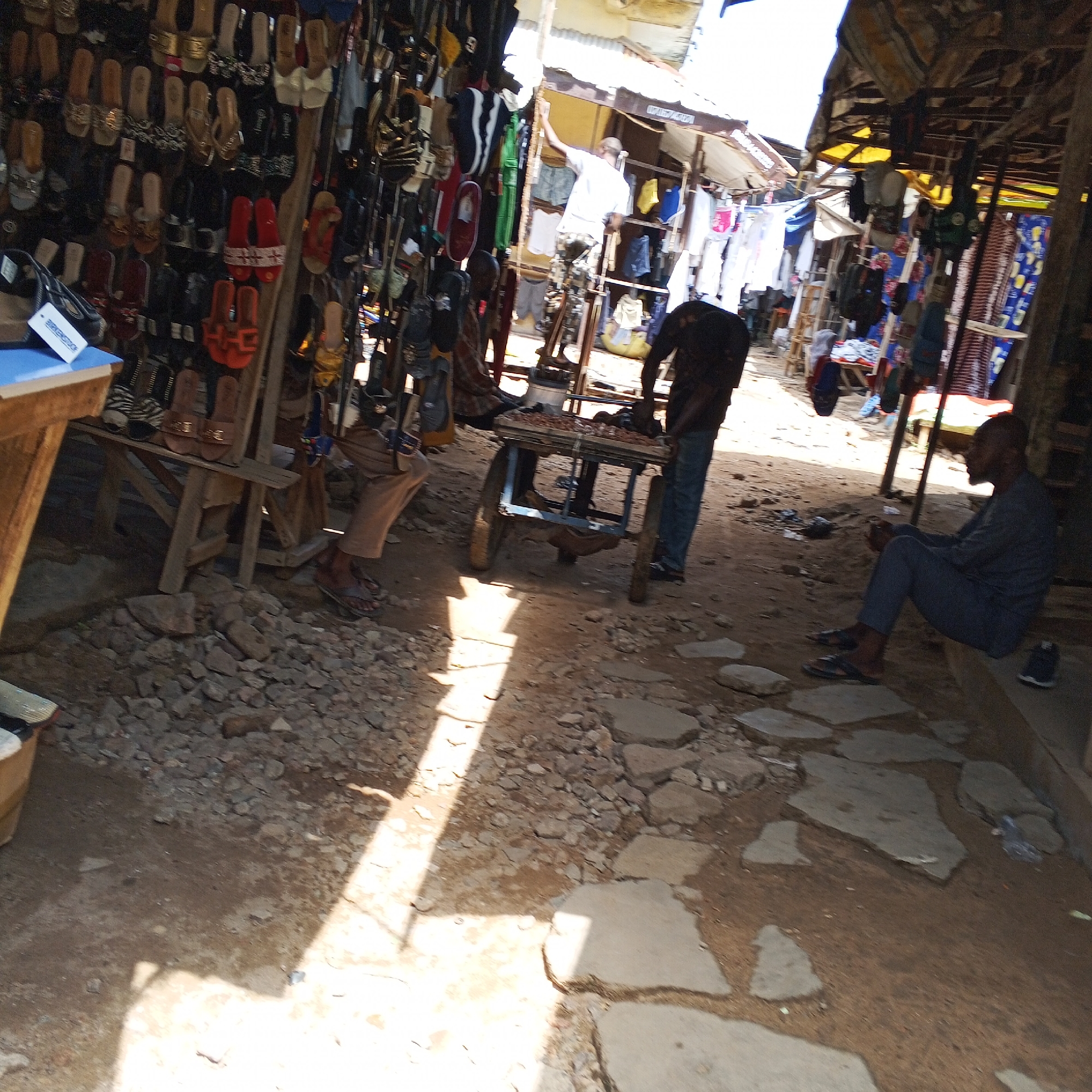 The image depicts an outdoor market scene, likely in Arawa, Nigeria, characterized by a bustling atmosphere and an array of merchandise. In the foreground, a man is seated on a low ledge, facing right, his hands clasped. To his left, a person in blue jeans and a dark shirt stands beside a wheeled cart laden with items. Further to the left, another individual, partially visible, appears to be in motion. The ground is covered with a mixture of dirt, small stones, and scattered flat stones.
The background reveals stalls with various goods, predominantly footwear, displayed on shelves and hanging from structures. Various clothing items are also visible, suggesting a diverse market. Sunlight streams in, casting strong shadows across the ground, indicating it is daytime. The overall impression is that of a vibrant, albeit somewhat cluttered, marketplace.