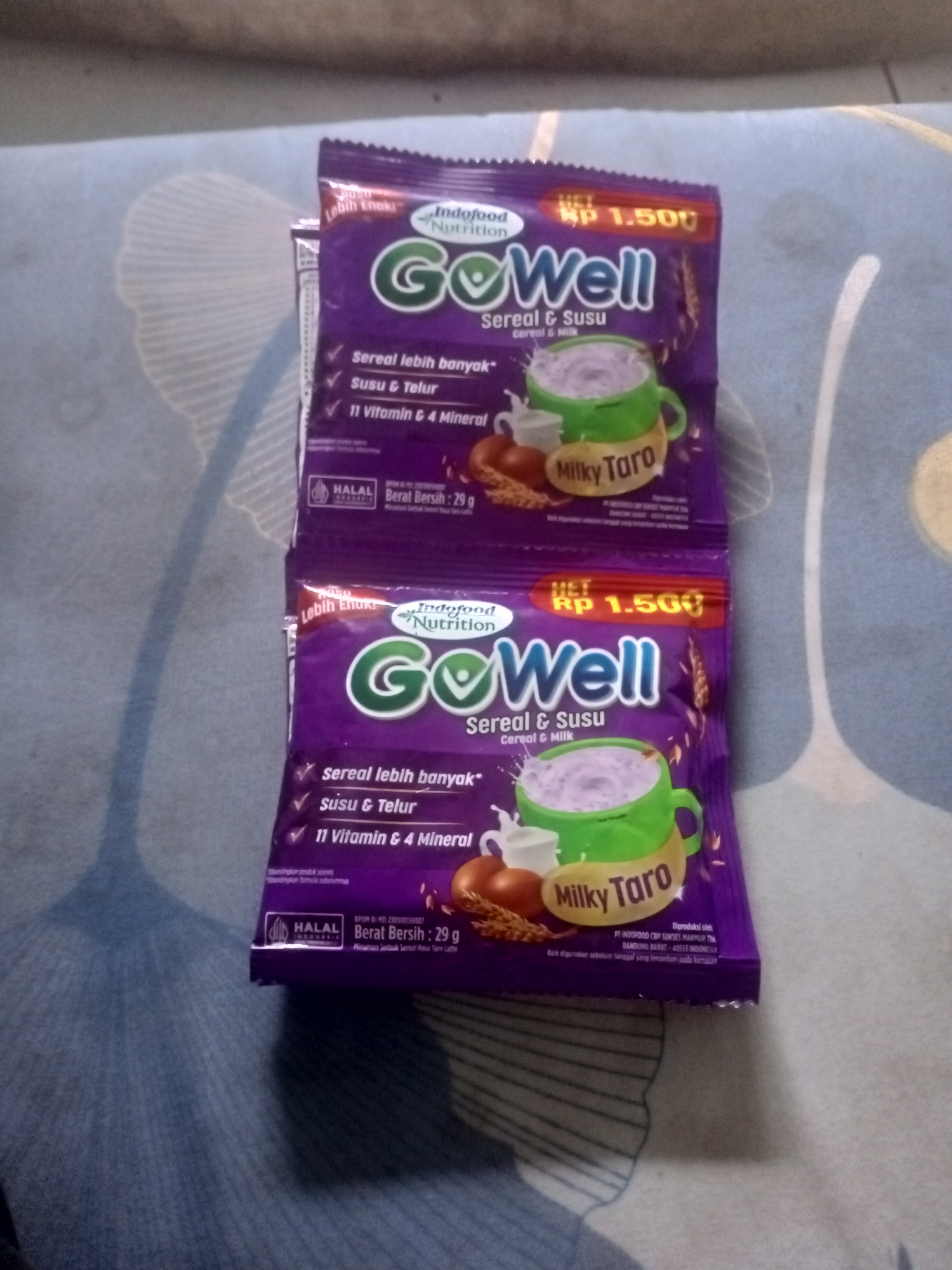 The image shows two purple GoWell cereal and milk sachets, one stacked on top of the other, with a portion of another sachet visible at the bottom. The sachets are branded with "Indofood Nutrition" and feature an illustration of a mug filled with purple liquid, a pitcher of milk, and a couple of eggs. The text on the sachets indicates they are "Sereal & Susu" (Cereal & Milk) with "Milky Taro" flavor. Both sachets display a price of "Rp 1.500". The lower sachet also shows "Halal Indonesia" certification and "Berat Bersih: 29 g" (Net Weight: 29 g). The background appears to be a blue and white patterned fabric, possibly a bedsheet. The image quality is blurry and the lighting is dim, suggesting it was taken indoors. There are no people or animals visible in the image. The scene does not provide any information about the location in the city of Badas, Indonesia.
