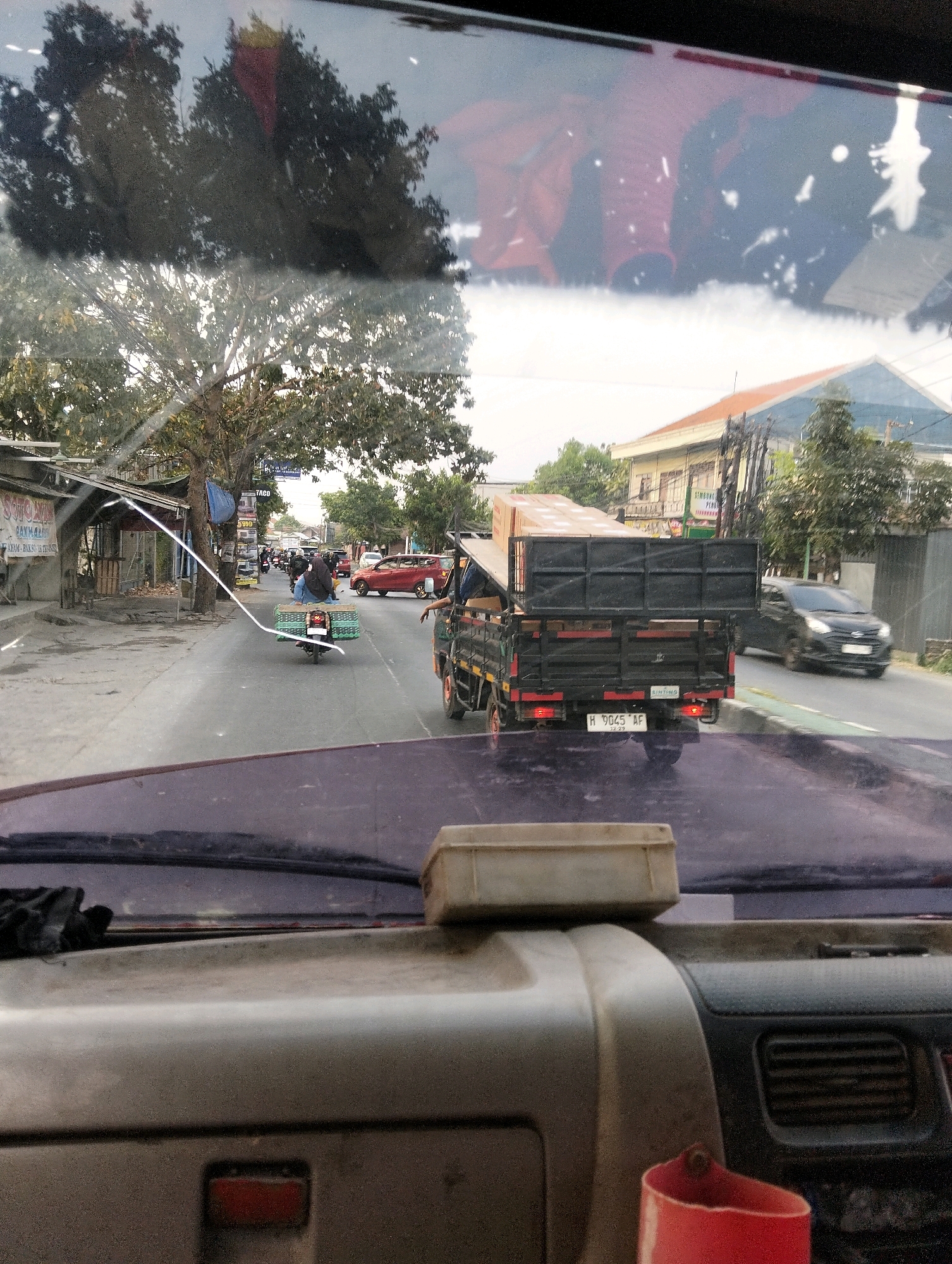 The image is taken from the perspective of a driver looking through a vehicle's windshield in Semarang, Indonesia. The view captures a street scene with vehicles and buildings.
In the foreground, the dashboard of the vehicle is visible, with a beige plastic container on top of it, and a red cylindrical object to its right.
The scene outside shows a two-lane road. Several vehicles are present: a black truck with a flatbed carrying wooden planks is in the right lane, moving away from the camera. Its license plate is visible and reads "H 9045 AF". A black car is in the left lane, also moving away from the camera. A motorcycle with a basket attached to the rear is in the left lane, in front of the truck and car. The rider of the motorcycle is wearing a headscarf. Further down the road, more vehicles and pedestrians are visible.
On the left side of the road, there are several small shops with open fronts. Trees line the side of the road, and power lines are visible. On the right side of the road, buildings with tiled roofs are present, along with more trees. The sky is overcast. The overall impression is of a busy street scene in an Indonesian city.