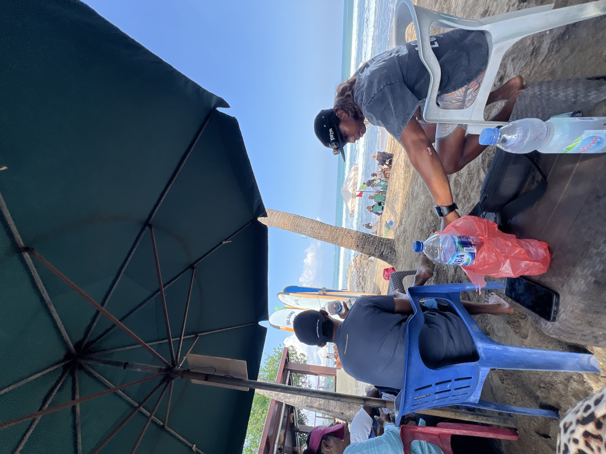 In Kuta, Indonesia, on a beach under a large, dark green umbrella, several individuals are seated on plastic chairs. The ground is covered in sand, and the ocean with moderate waves is visible in the background, under a clear blue sky. Palm trees are present near the seating area.
In the foreground, a person in a black baseball cap and dark t-shirt is seated on a white plastic chair, leaning forward. A black bag is on the sand in front of them, and two plastic bottles of water and a red plastic bag are on a low table next to the chair. A cell phone is also on the table. To the left, a person in a dark blue t-shirt and a black baseball cap is seated on a blue plastic chair, holding a bottle of water. Another person, wearing a pink visor and a light blue top, is partially visible on the far left. Further back on the beach, several other people are scattered around, some sitting on chairs under umbrellas, and some in the distance near the water. Surfboards are propped up against a structure in the middle ground.