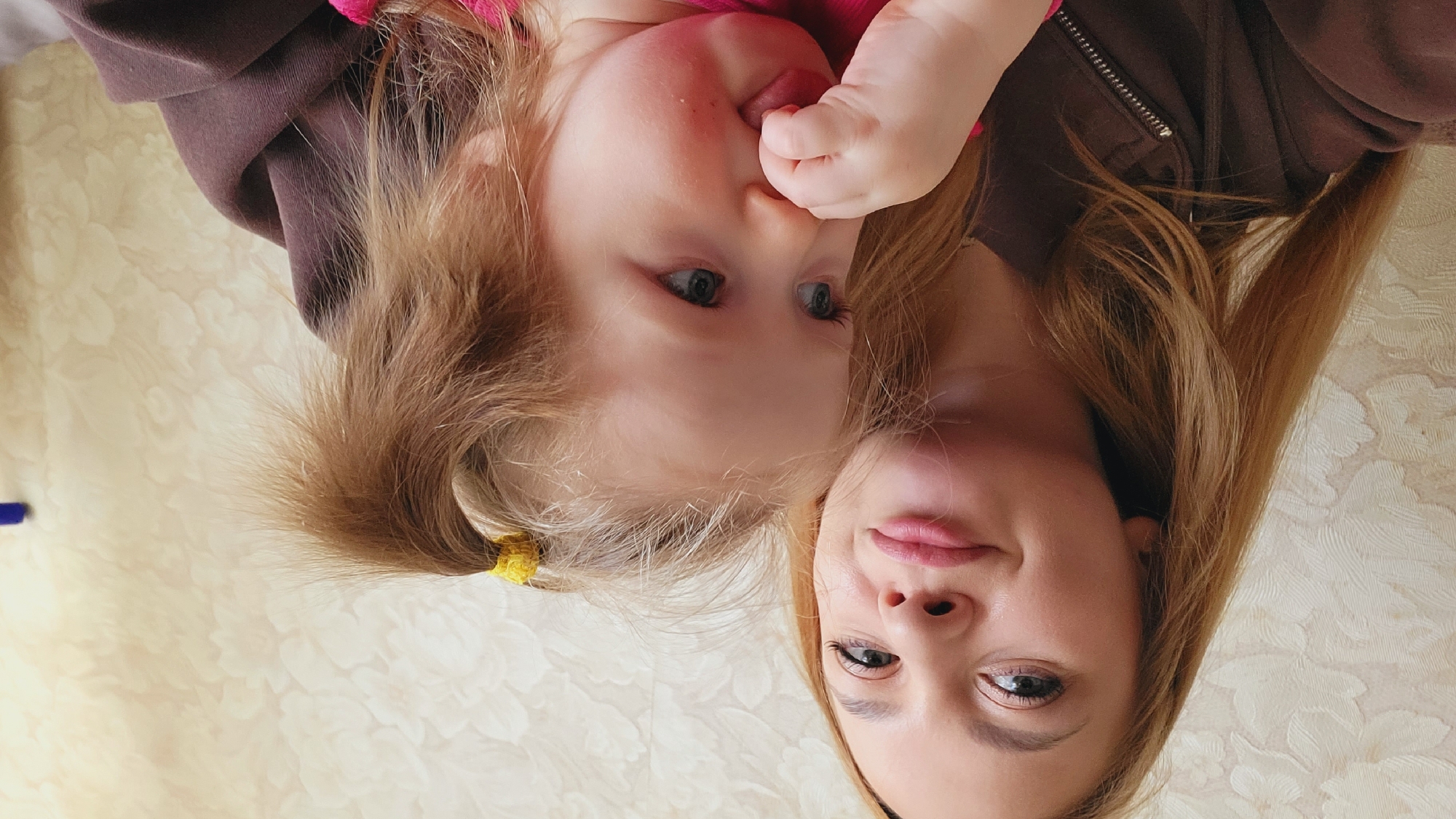 A photograph depicts an adult and child, likely mother and daughter, in an indoor setting. The image is rotated; the woman's face is positioned in the lower half, and the child's face in the upper half. The child has light blonde hair pulled back in a small ponytail with a yellow hair tie, while the adult has straight, light brown hair. The child's hand is raised to her mouth, with her finger possibly in her mouth. The adult is looking towards the camera. The backdrop is a light-colored wall with a floral pattern. Based on the request for the location, it is likely that this scene is in the city of Chaikovsky, Russia.