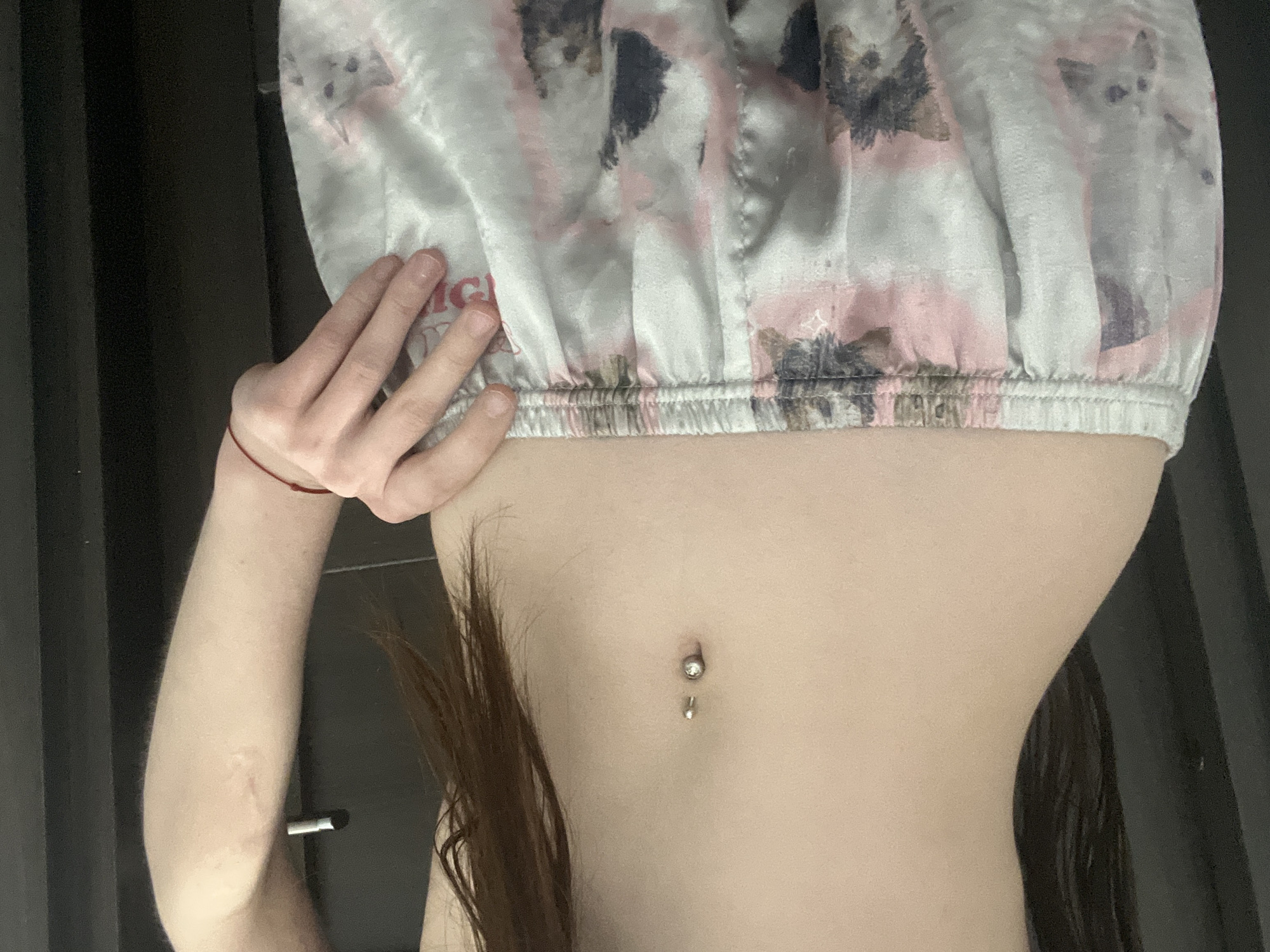 The media file presents a medium-close-up image of a person's upper torso, arm, and hand, rotated 90 degrees clockwise. The scene is set indoors, against a dark background that resembles curtains or wall panels.
The primary subject is a fair-skinned individual, likely female, whose midriff is prominently displayed. They are wearing a light-colored, possibly silky, crop top or pajama top with an elasticated bottom hem. The clothing features an abstract pattern of pink, grey, and dark mottled shapes, which could be interpreted as stylized animals like cats or foxes, or simply a decorative design. Partially visible text on the clothing appears to read "MG" followed by "WO" or "WOR".
A silver belly button piercing with a small dangling charm is visible on the navel. The person's left arm is raised, with the hand gently resting on the bottom edge of their top. A thin red string is tied around the wrist. A subtle, lighter-colored scar is discernible on the inner forearm. Long, dark brown hair cascades down the left side of the torso.
The activity taking place appears to be posing for a photograph, possibly a selfie, highlighting the midriff, clothing, and piercing. No specific time of day is discernible, but the setting is clearly indoors. The image provides no visual cues for its location, but the context provided is Saint Petersburg, Russia.