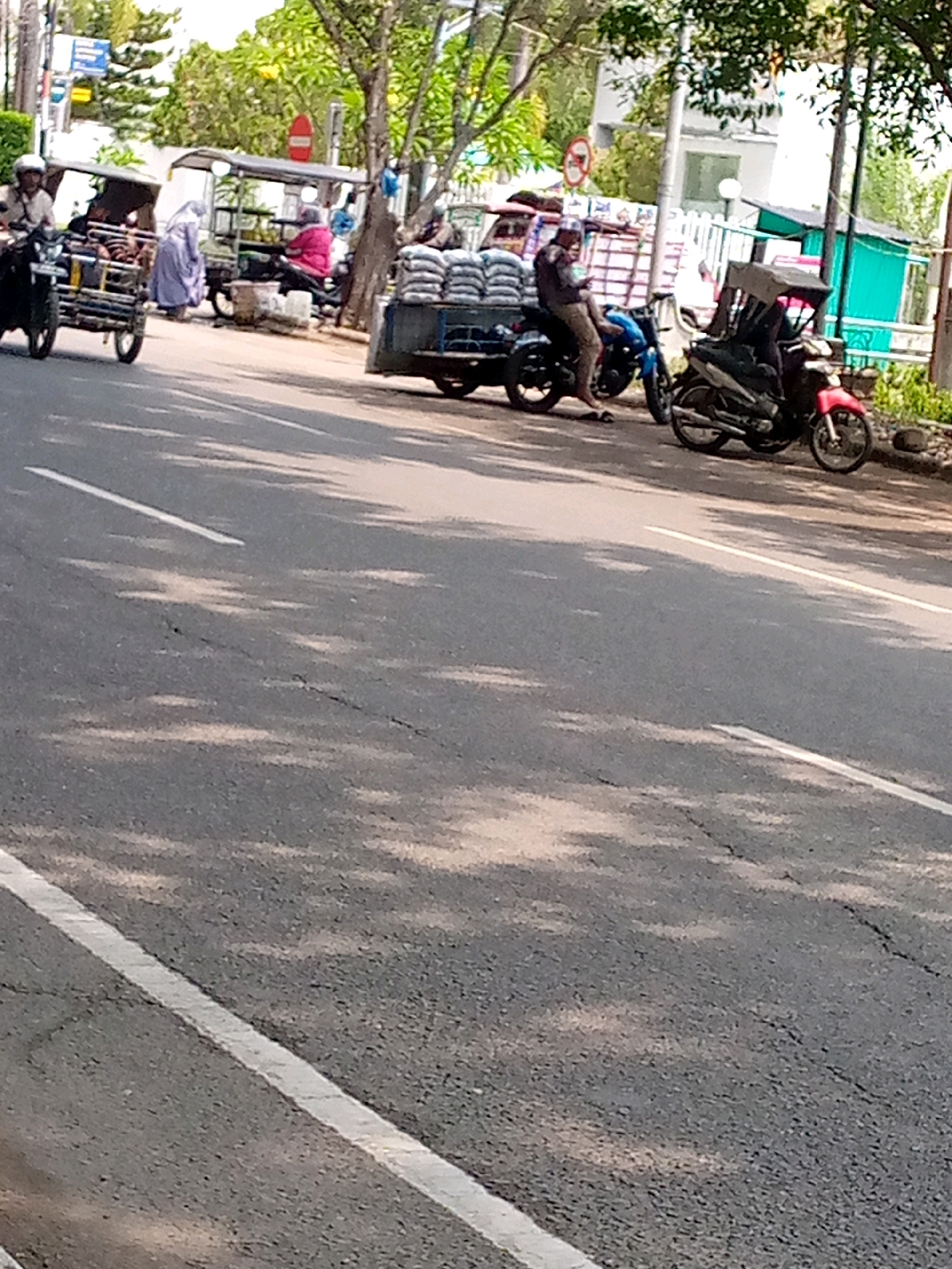 The image depicts a street scene in Lhokseumawe, Indonesia, with several people and vehicles. In the foreground, a gray asphalt road with white lane markings is visible. In the background, a street vendor appears to be selling goods from a cart. A man is seated on a motorcycle, and another motorcycle is parked nearby. Two individuals are standing next to a cart loaded with sacks of what appear to be rice. One person is wearing a light purple abaya and a headscarf, while another is wearing a pink headscarf. The environment suggests a commercial or market area, with trees and buildings visible in the distance. The overall scene is bustling with activity, indicating a typical day in a public area.
