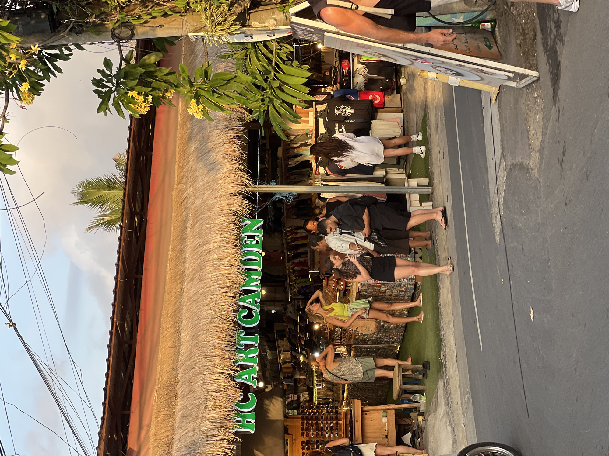The image depicts a street scene in Kuta Selatan, Indonesia, in front of a shop with a thatched roof and a sign reading "HC ART CAMDEN." The shop appears to sell clothing, as racks of garments are visible inside. Several people are standing outside the shop, some browsing and some talking. The lighting suggests it is either early morning or late afternoon, with warm, soft light illuminating the scene. The street in front of the shop is paved, with a white line marking the center. There are power lines overhead, and a palm tree is visible in the background. A person on the right side of the image is partially visible, appearing to be holding a sign. The overall atmosphere is casual and relaxed, typical of a tourist area.