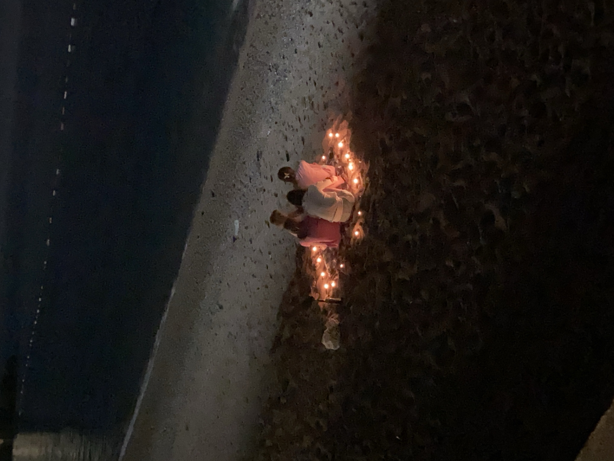 Three people are kneeling near lit candles on the ground in Petrovac centar, Montenegro. The setting is dark, likely at night, and the ground appears to be rocky or pebbled. A road or wall runs along the left edge of the frame.