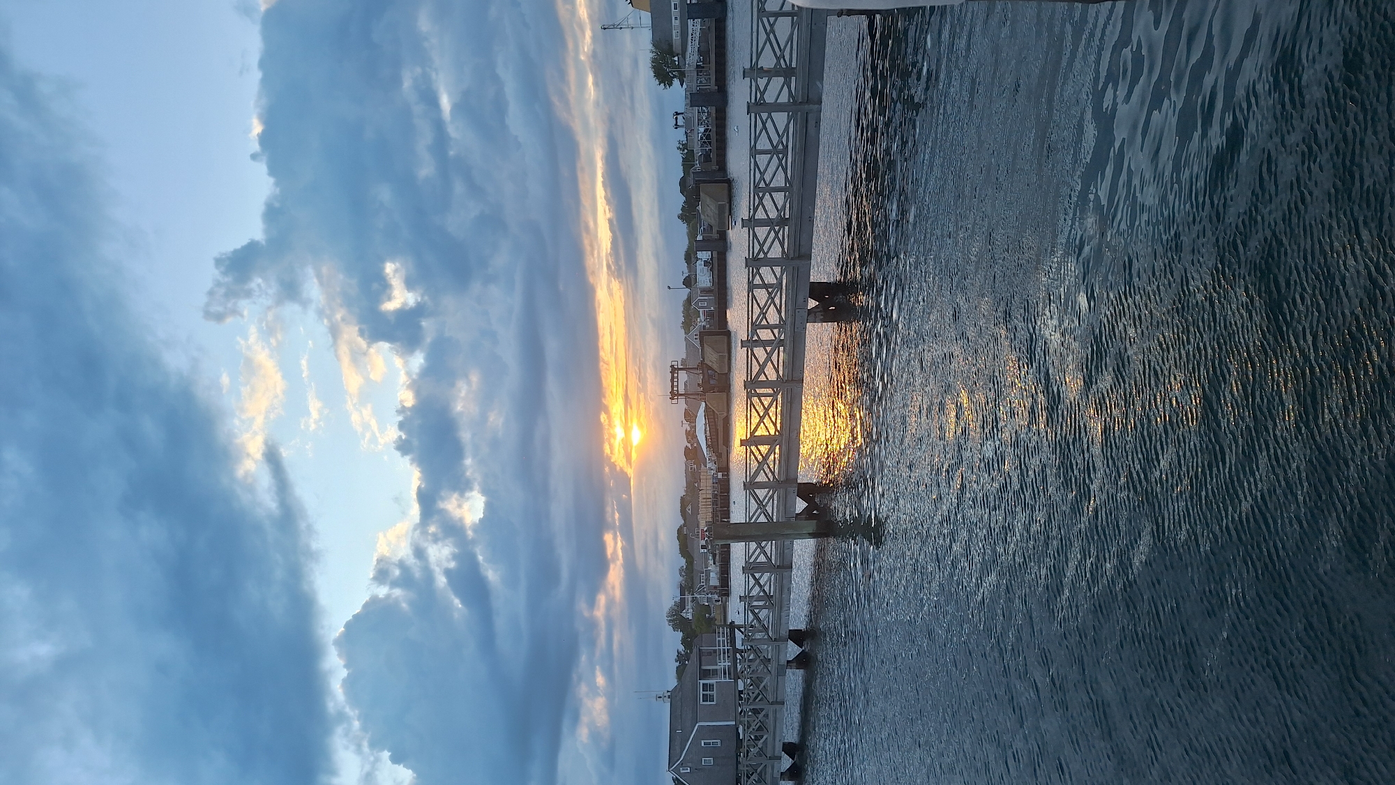 This image depicts a waterfront scene in Nantucket, Massachusetts, USA. The sun is setting, casting a warm glow on the water and the sky. A wooden pier extends into the water, with buildings and a ferry terminal visible in the background. The sky is filled with dramatic clouds, tinged with orange and yellow from the setting sun. The water is calm, with gentle ripples reflecting the light. The overall impression is one of peacefulness and natural beauty.