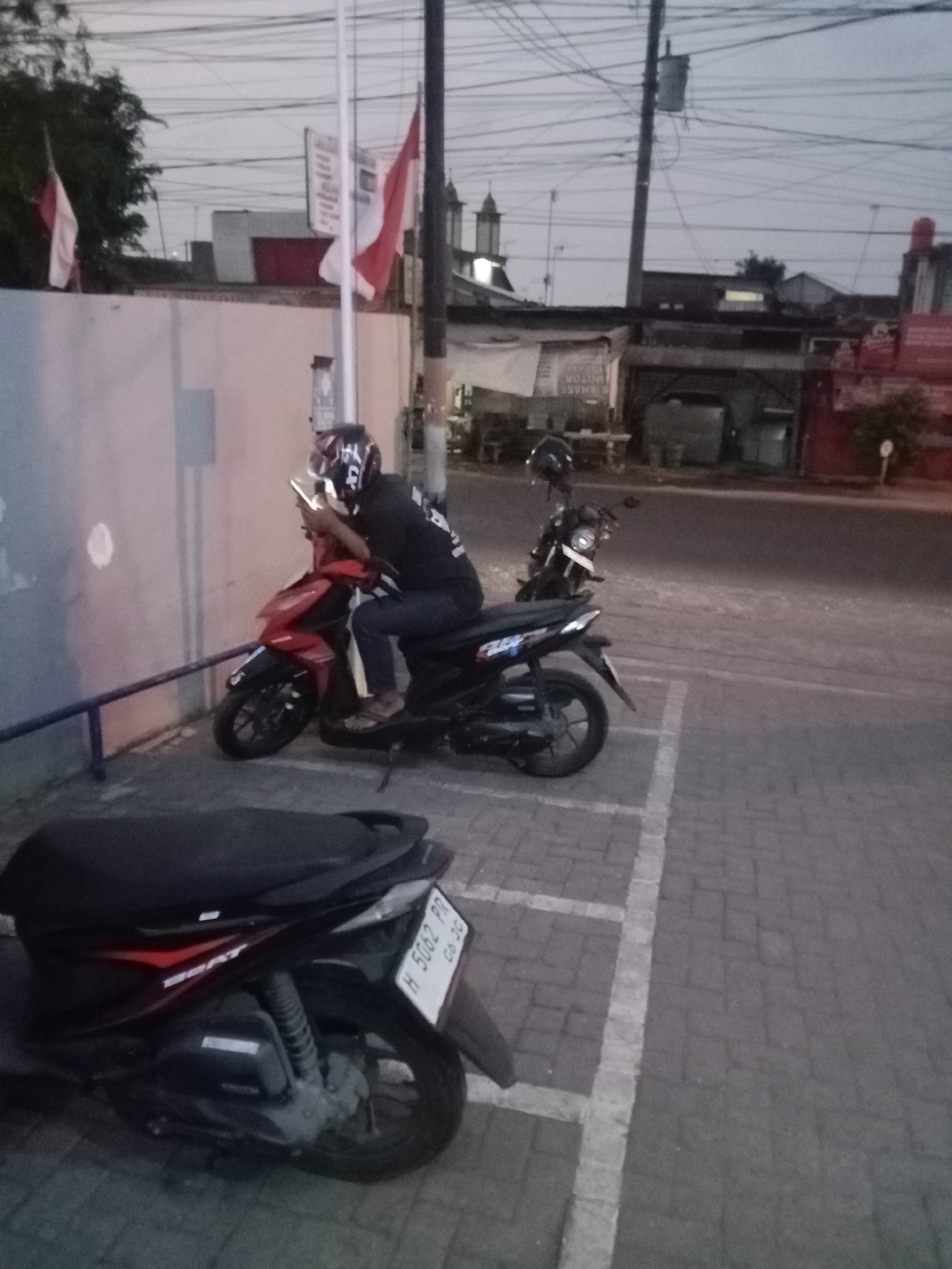 The image depicts an outdoor scene in Kramat, Indonesia, likely during dusk, given the ambient lighting and overcast sky. In the foreground, a dark-colored scooter is parked, its license plate reading "H 5062 PD 06-26". To the left of this scooter, a man wearing a helmet and a black shirt is seated on another red and black scooter. He is holding a smartphone in his hands and appears to be looking at it. The scooter he is on is parked next to a light-colored wall. Behind the man and his scooter, a second scooter is parked, facing away from the camera. In the background, buildings and various electrical wires are visible. Two Indonesian flags, red and white, are displayed prominently on poles in the middle ground, near a building with architectural elements resembling a church steeple. The ground is paved with interlocking bricks, and white lines demarcate parking spaces.