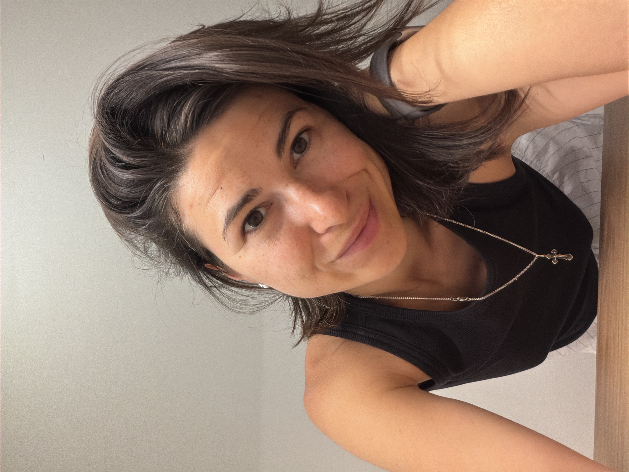 A woman with dark hair and brown eyes is looking at the camera. She is wearing a black tank top and a silver necklace with a cross pendant. She has a grey smartwatch on her wrist. The background is a plain grey wall. The location is unspecified and unknown.