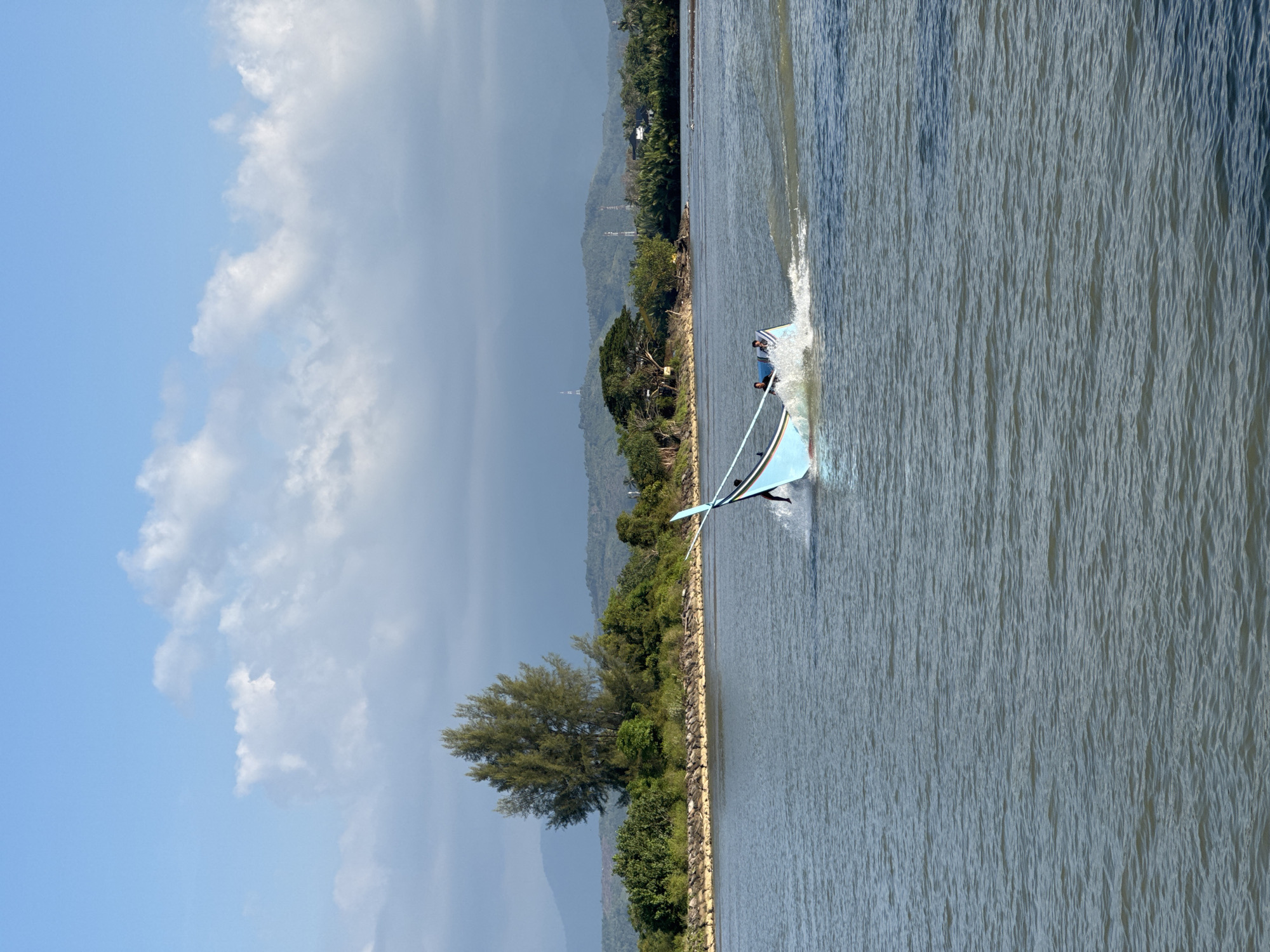 A bright blue and white motorized fishing boat with two occupants is moving at speed through a body of water, creating a significant wake. The boat has a pointed bow and a flat stern. The water is a murky grey-blue with small ripples. In the background, a narrow strip of land is densely covered with green foliage, including a tall, spindly tree. Beyond the trees, rolling hills are visible under a bright blue sky with scattered white clouds. The scene suggests a coastal or riverine environment in a tropical location. The location is identified as Reudeuep, Indonesia.
