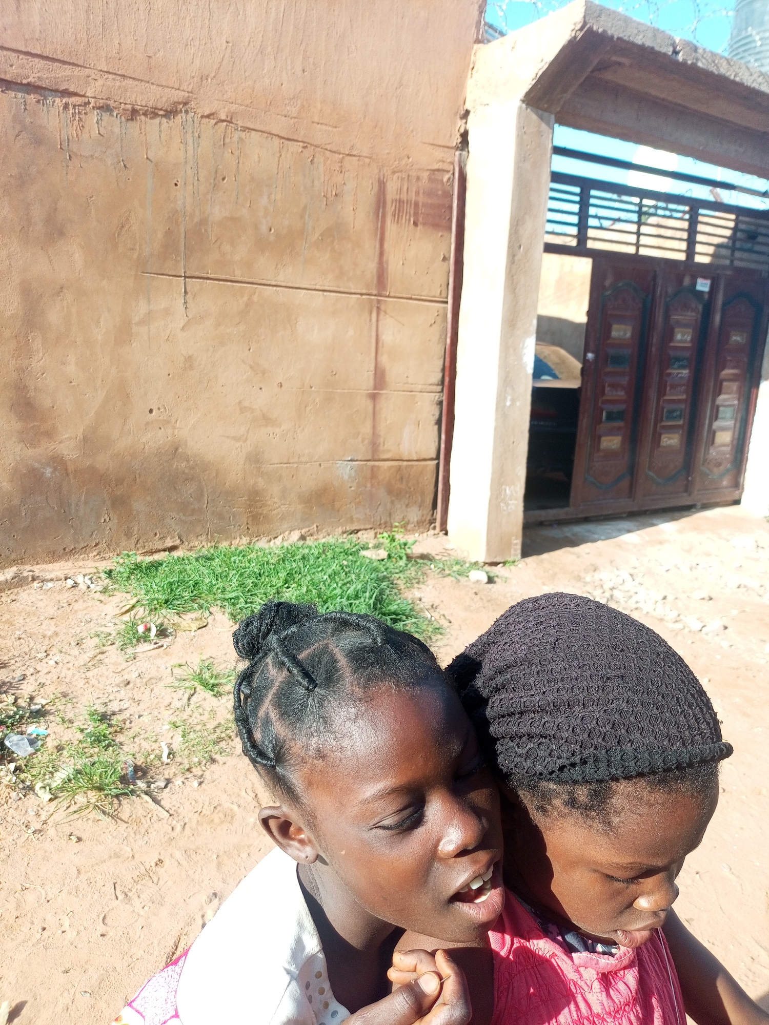 In Tempure, Nigeria, two young girls are captured in a close-up shot against a backdrop of a weathered, sandy-brown wall. The wall exhibits vertical streaks and horizontal indentations. To the right of the wall, an archway frames a set of dark brown, ornate double doors with rectangular panels. Sunlight casts shadows across the scene, illuminating patches of dry grass and scattered debris on the sandy ground. The girl on the left, facing forward and slightly to the right, has her eyes closed and mouth open, with intricate braids in her hair. She is wearing a white top. Beside her, the girl on the right, wearing a black knitted cap and a pink garment, leans in, her face angled downwards, with her eyes partially open. Their bodies are close, suggesting an intimate interaction. The overall impression is one of a candid, outdoor moment in a sun-drenched, rustic environment.