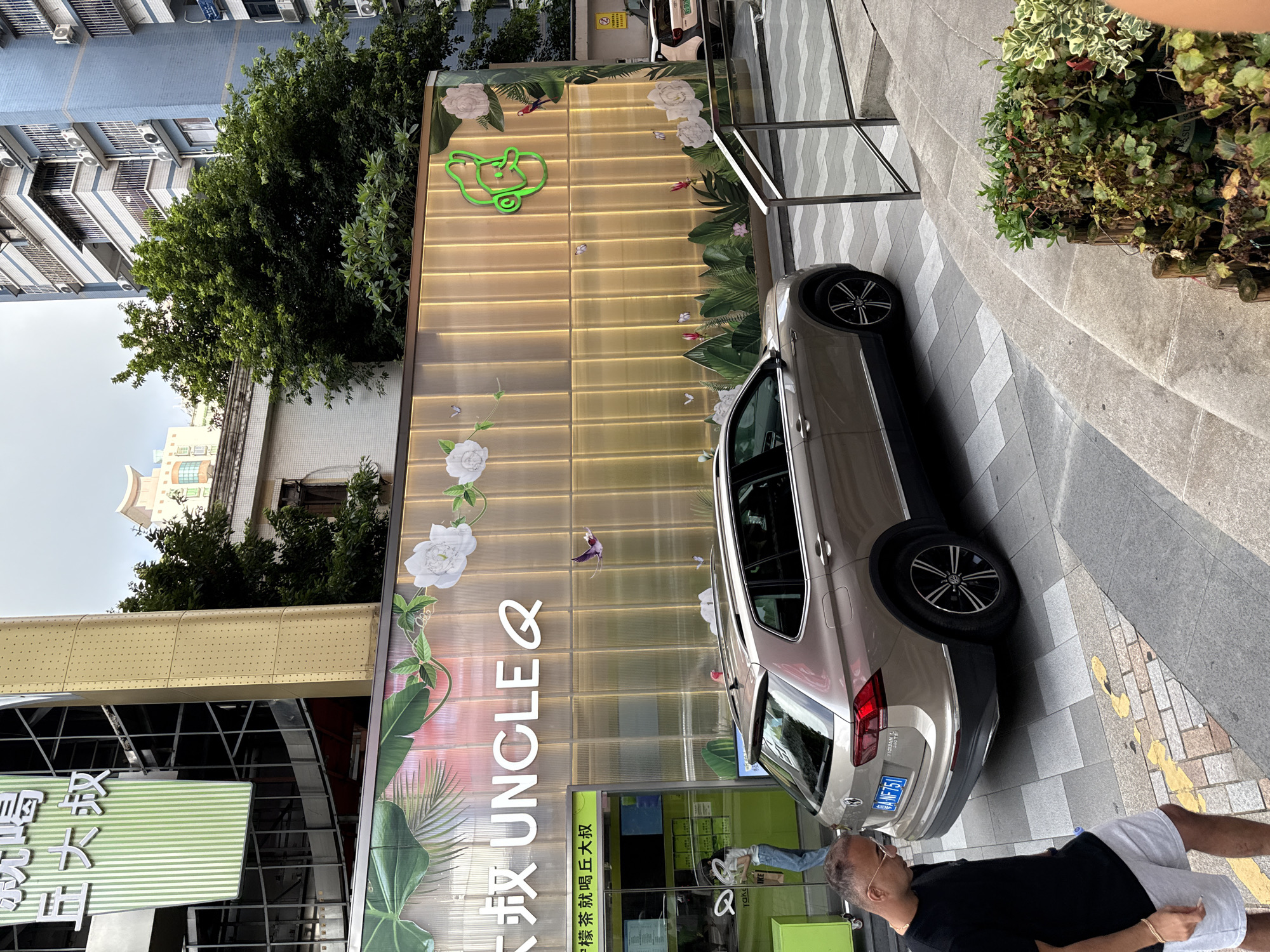 In Haizhu District, China, a brown Volkswagen Tiguan is parked on a street in front of a building with a distinctive storefront. The building's facade is adorned with a mural featuring large white flowers, green foliage, and several colorful birds and butterflies. The words "UNCLE Q" are prominently displayed in white lettering against a translucent, ribbed background that is illuminated from behind with a warm, golden light. To the left of the storefront is a bright green booth with a menu visible.
A man wearing a black t-shirt and light grey shorts walks away from the camera, looking down. Another person, a woman in a white shirt and blue jeans, is seen inside the green booth, holding a bag. A second vehicle, a light-colored SUV, is partially visible further down the street. The sidewalk features a decorative pattern of grey and white tiles, and a curved stone staircase leads down to a planter box filled with greenery. The background shows a blue apartment building and trees.