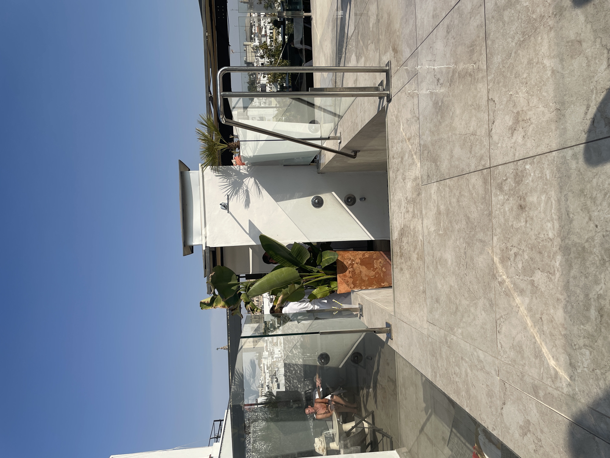 This is a medium shot, taken from a slightly elevated outdoor perspective, looking across a tiled patio and down a short set of stairs. The scene appears to be on a rooftop in Seville, Spain, given the distant cityscape with distinctive architecture. The sky is a clear, vibrant blue.
In the foreground, a light-colored, marble-patterned tiled patio leads down a short flight of concrete stairs. A modern metal and glass railing system is installed along the stairs and on the patio.
To the left, a glass-enclosed area reveals an indoor space. Inside, a woman with tan skin is seated at a small round table, wearing a patterned swimsuit. Another person is partially visible behind her. A chair and what appears to be a bag are also present in this area.
Further back, a large potted plant with broad, green leaves, possibly a Bird of Paradise, stands in a rustic, orange-toned pot. Behind the plant, two individuals are visible. One, wearing a white robe, is standing and appears to be interacting with the plant or something near it. Another person, wearing a light-colored shirt, is partially obscured. A white wall with shadows from palm fronds is visible behind them, and a shower fixture is mounted on this wall.
The background displays a cityscape typical of Seville, with white buildings and red-tiled roofs, suggesting a sunny, warm day. The overall impression is of a modern rooftop terrace overlooking a historic Spanish city.