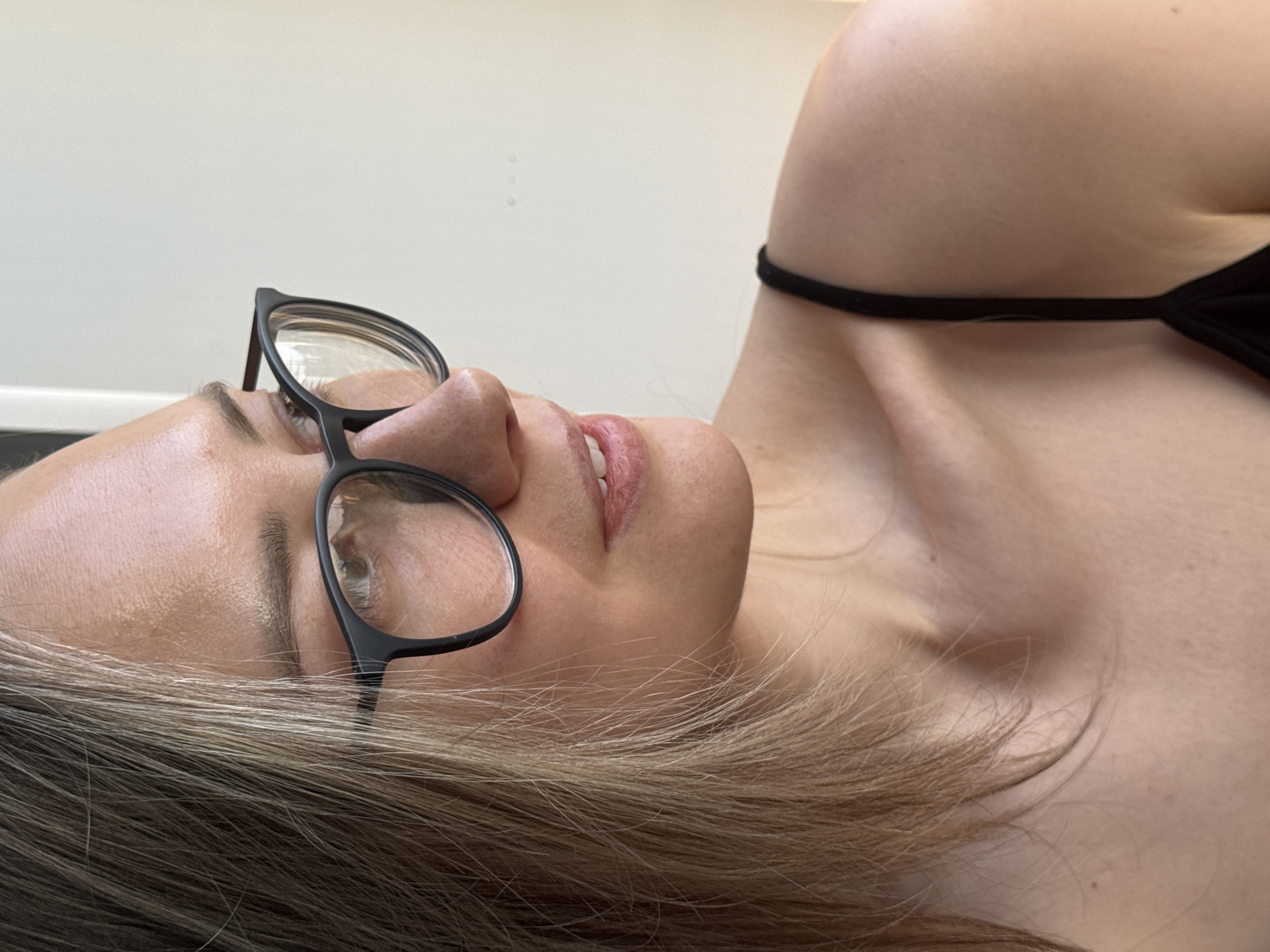 A woman with blonde hair and glasses lies on her back. She wears a black garment that partially covers her upper chest. The background is a plain white wall and a dark surface. There is no information to suggest that the image was taken in Chaikovsky, Russia.