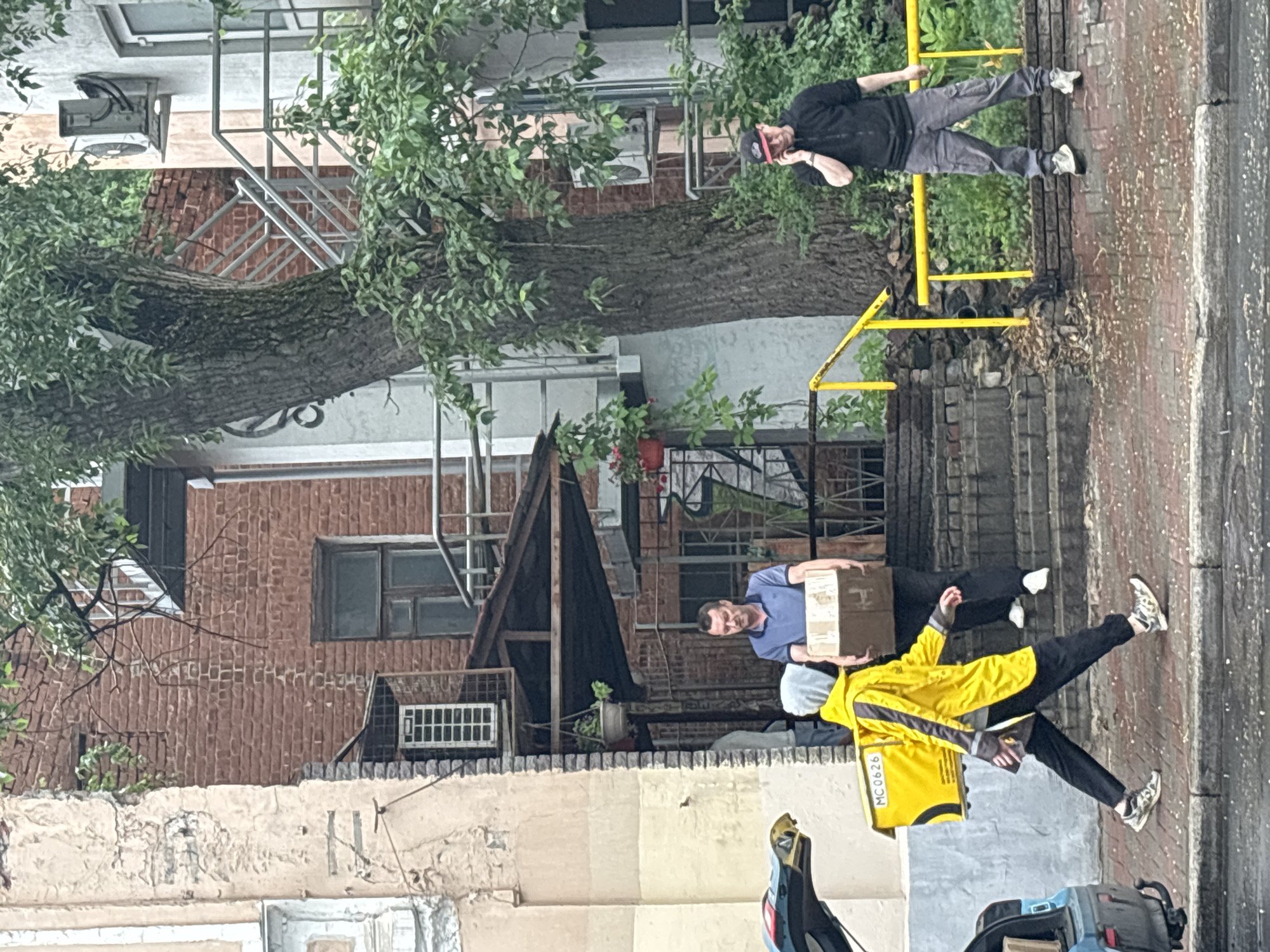 In an urban setting in Nizhny Novgorod, Russia, on a brick and stucco building facade, a man in a blue polo shirt stands on a set of concrete stairs, holding a brown cardboard box. To his left, a person wearing a bright yellow hooded jacket and black pants, with a yellow delivery box on their back labeled "MC0626," walks across a brick sidewalk. To the right, a man in a black hooded jacket and gray cargo pants stands on a yellow railing, talking on his mobile phone. A large tree with green foliage is visible to the left of the stairs, and a car with its trunk open is parked on the left side of the image. The ground appears wet, indicating recent rain.