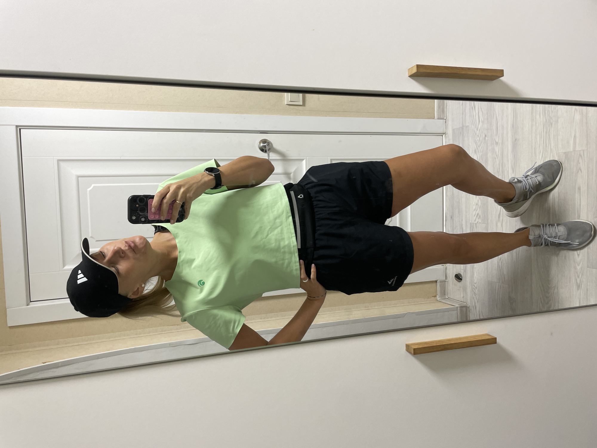 A woman is taking a mirror selfie. She is wearing a black Adidas baseball cap, a light green t-shirt, black shorts, and gray sneakers. She is holding a phone in her left hand. The reflection in the mirror shows a white door with a silver doorknob, light beige wall, and gray laminate flooring. Two small wooden shelves are affixed to the wall above and beside the mirror. The city of Baranovka, Russia is not discernible from the image.