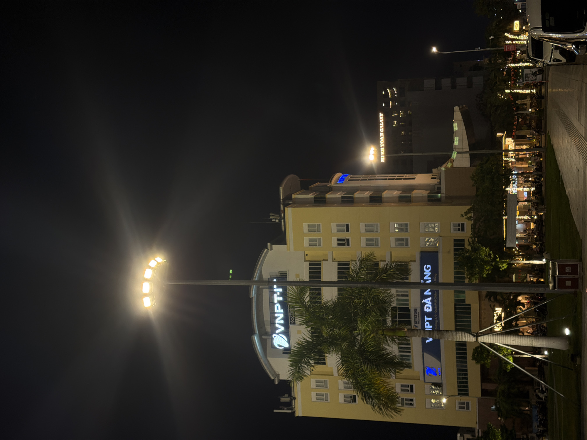 The image captures a nighttime scene in Đà Nẵng, Vietnam, focusing on a brightly lit yellow building with a large "VNPT-I" sign on its upper facade. A palm tree stands in front of the building, its base illuminated by spotlights. To the right, another building is visible with the text "MISH TOAN GALAXY" illuminated on its exterior. The foreground shows a paved walkway, a grassy area, and several parked motorcycles. The scene is further enhanced by decorative string lights and streetlights, creating a lively urban atmosphere.