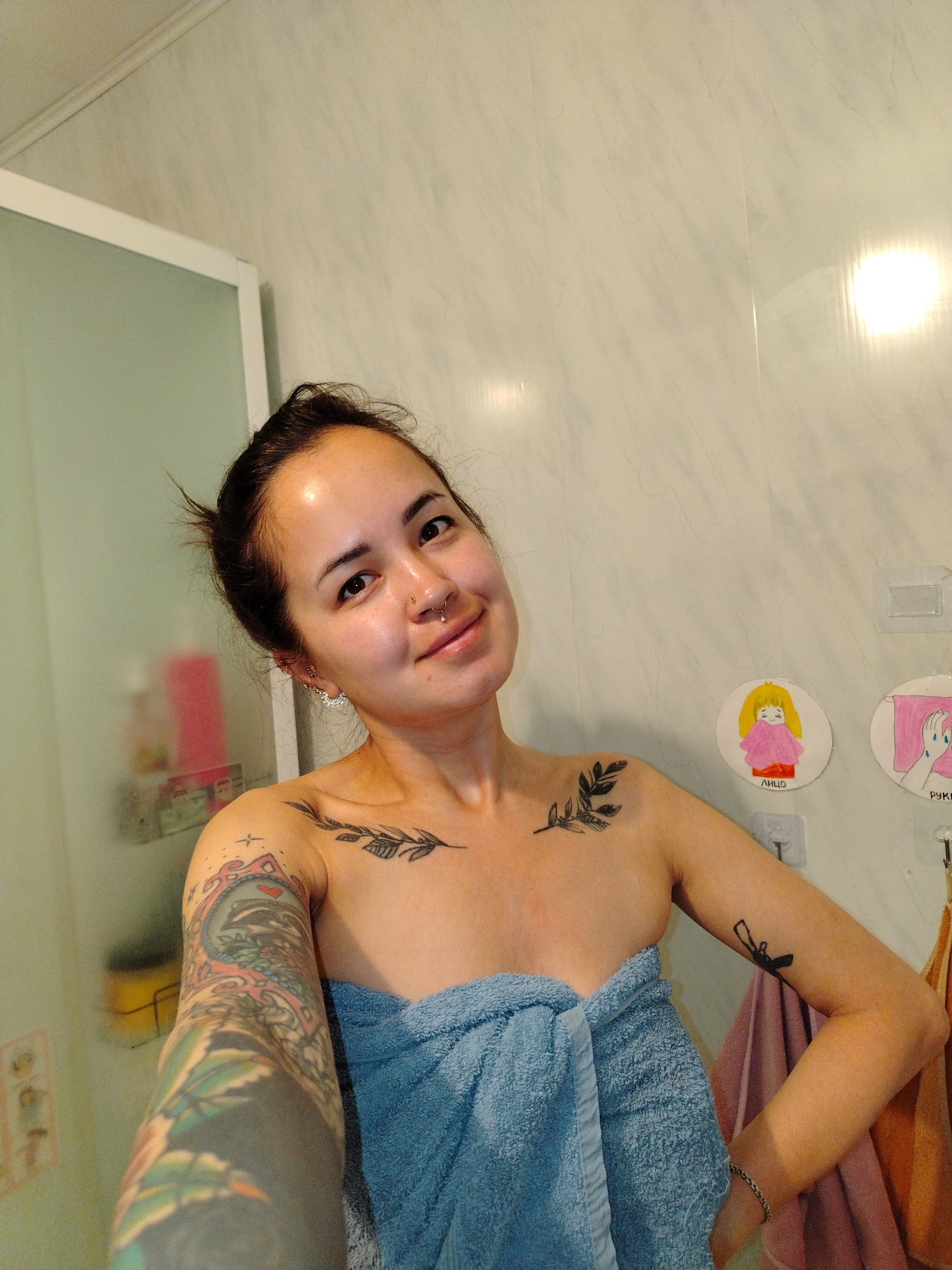 A woman is in a bathroom, wrapped in a light blue towel. She has dark hair pulled back and a nose ring. Visible tattoos include a sleeve on her left arm and leaf-like designs on her chest. The bathroom walls are light colored, and there are illustrations hung on the wall. A shower door and various bottles are visible in the background.