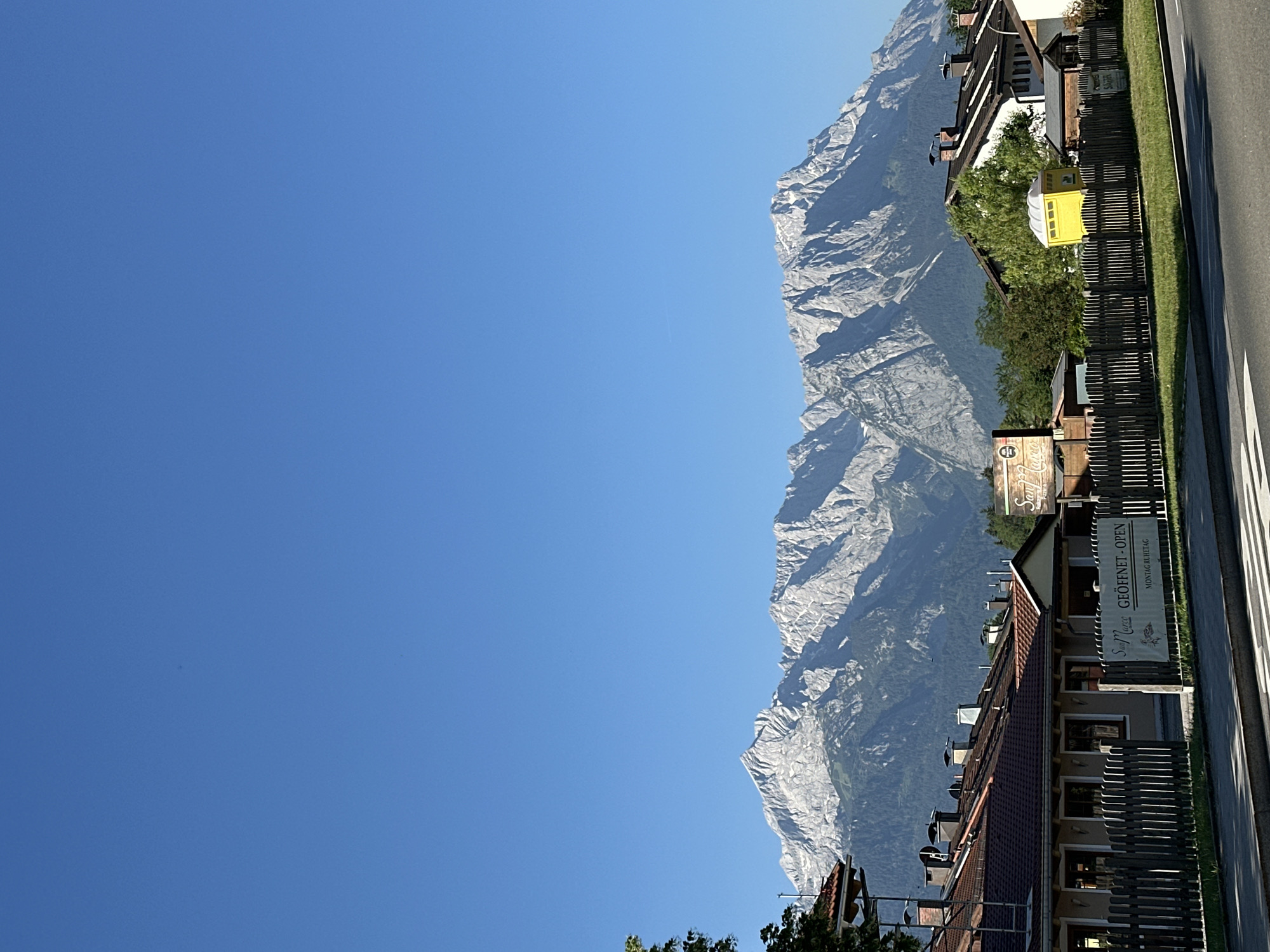 The image shows a picturesque view of the Alps in Garmisch-Partenkirchen, Germany. In the foreground, there are several buildings with red tiled roofs, a wooden fence, and a paved road with white markings. A yellow portable toilet is visible behind the fence. Behind the buildings, the majestic Alps mountain range dominates the background, with its rugged peaks and snow-capped summits. The sky is a clear, vibrant blue, indicating a sunny day. The overall scene conveys a sense of natural beauty and tranquility.