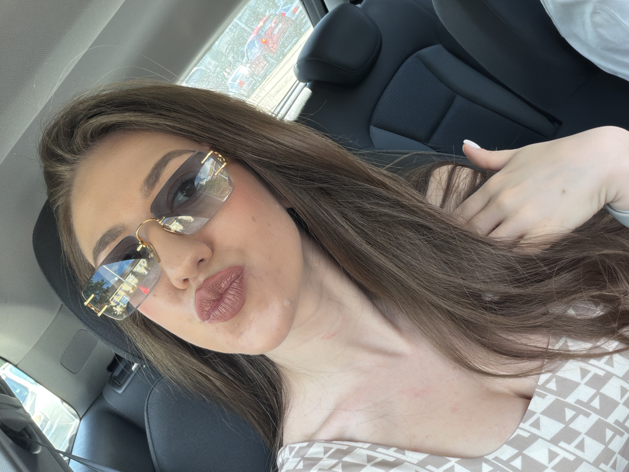 A young woman with long brown hair is photographed from the chest up, inside a car. She is wearing oversized sunglasses with gold frames and has on a light-colored top with a geometric pattern. Her lips are pursed, and she is looking at the camera. The background shows the interior of the car, including black seats and the window, through which other cars and buildings can be seen. The scene appears to be in a city, possibly Sofia, Bulgaria, given the style of the cars and architecture visible through the window. The lighting suggests it is daytime.