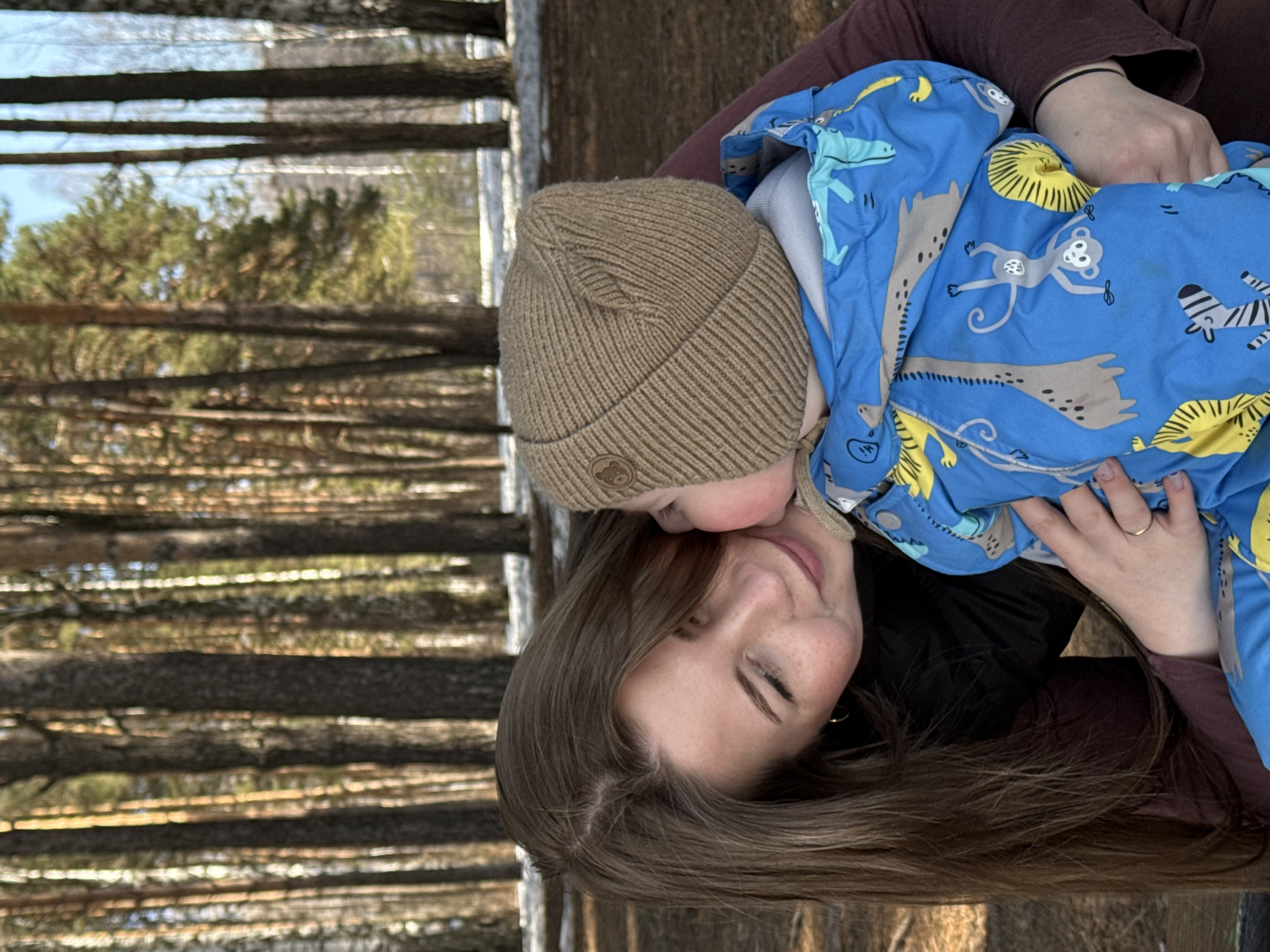 A photograph shows a woman holding a small child outdoors in Chaikovsky, Russia. The woman has light skin, long brown hair, and is wearing a maroon-colored long-sleeved shirt. She is smiling with her eyes closed as the child leans their face against hers. The child wears a brown knit hat and a blue jacket with animal print. The background consists of a blurred forest scene with many bare trees.