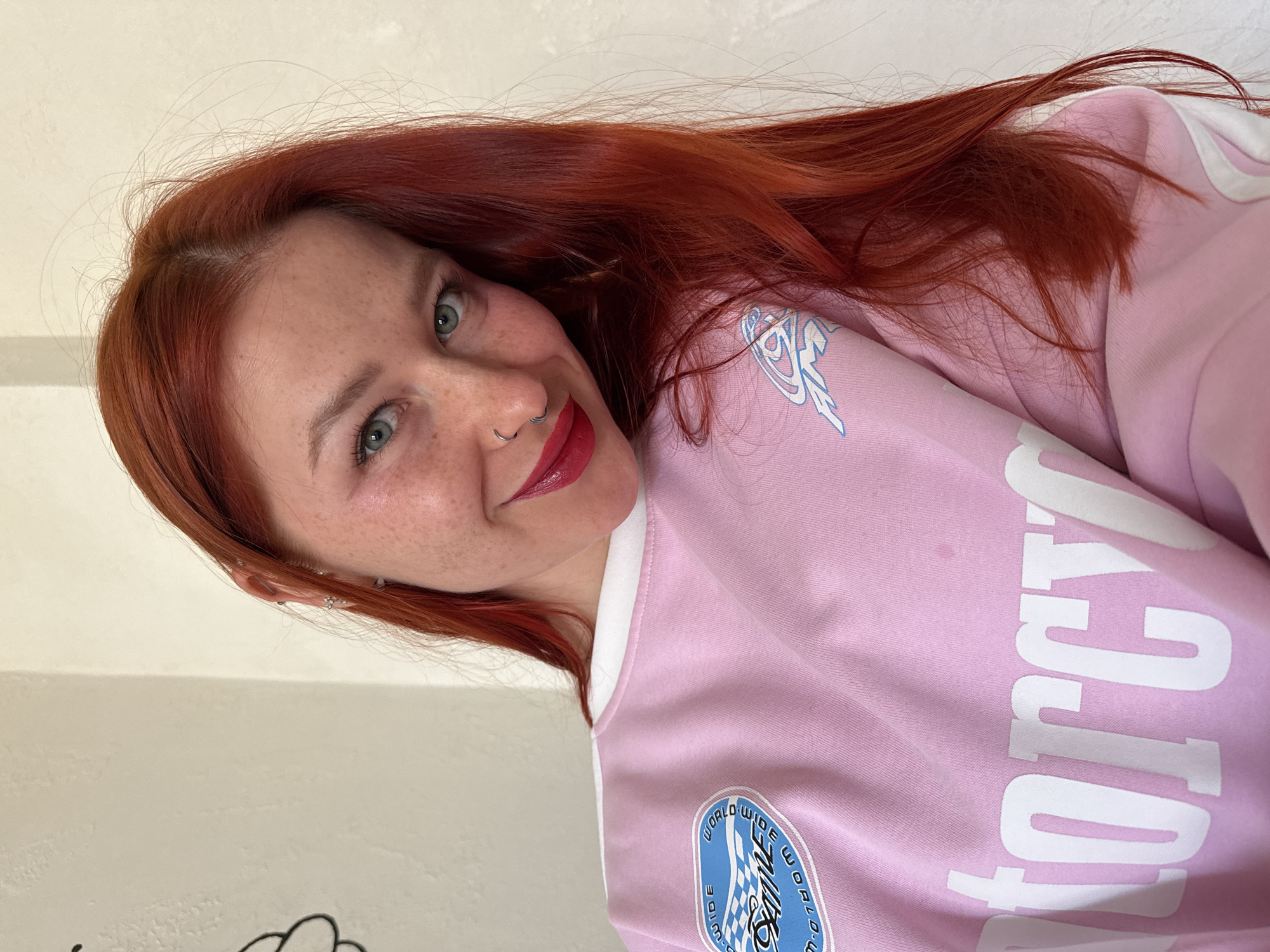 A fair-skinned woman with red hair and freckles is looking at the camera with a slight smile. She has a nose ring and is wearing red lipstick. The woman is wearing a light pink jacket with white trim and graphics. The jacket has a round logo on the front with blue and white details, and large white letters on the sleeve. The background is a plain, light-colored wall. The image is rotated slightly. The location is not evident from the image.