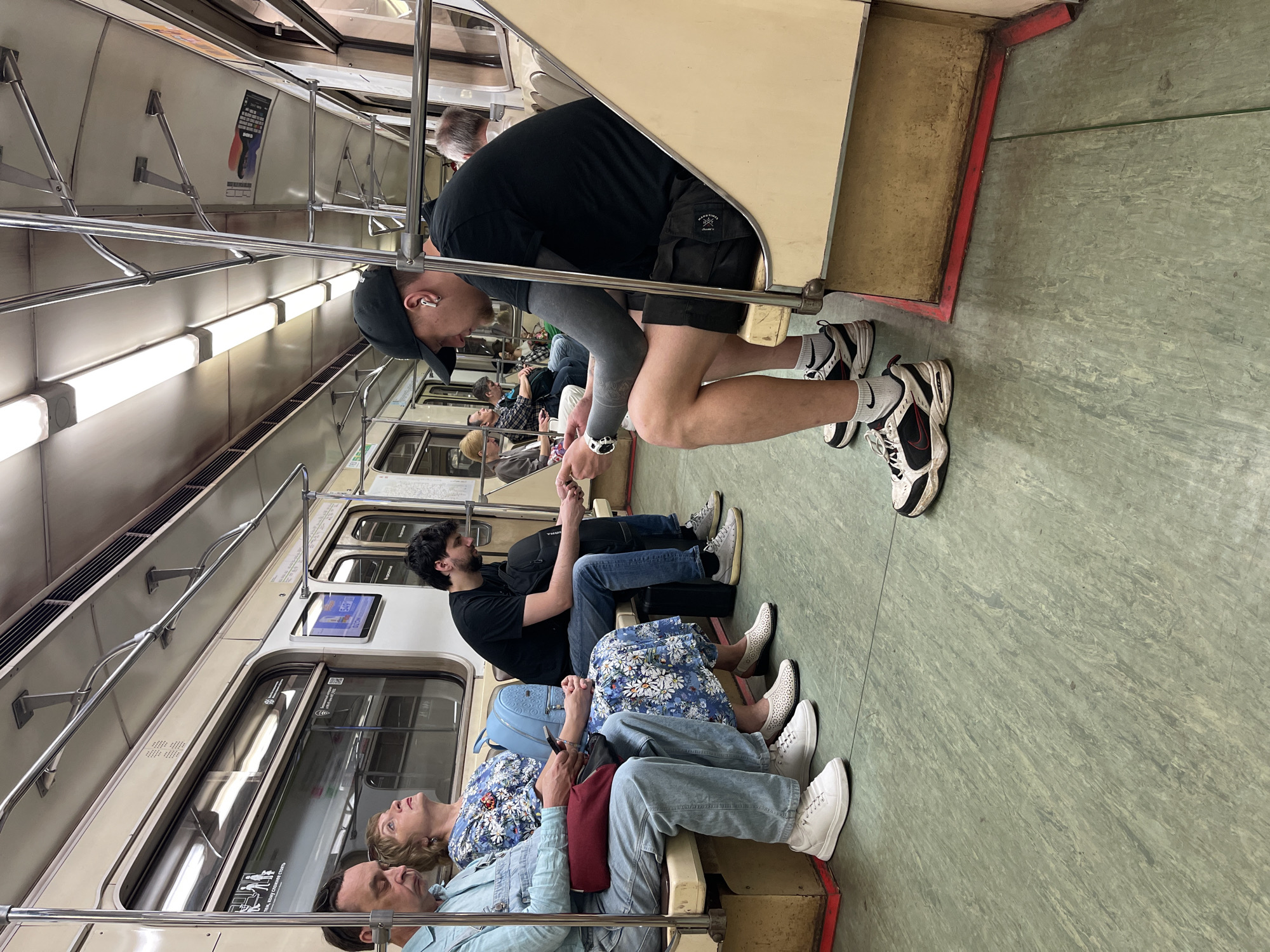 A subway car interior in an unidentified location shows multiple seated passengers and one standing male. The seated passengers, from foreground to background, include a male and female pair wearing light-colored clothing, a female in a blue floral dress, and a male in black. The standing male, wearing a black shirt, shorts, and baseball cap, leans forward with hands positioned in front, an electronic device on his wrist, and wireless earbuds in his ears. Flooring is green, and the walls are primarily light tan. Overhead lighting is fluorescent. Metal bars and fixtures line the subway car. A digital screen is visible above one of the windows.