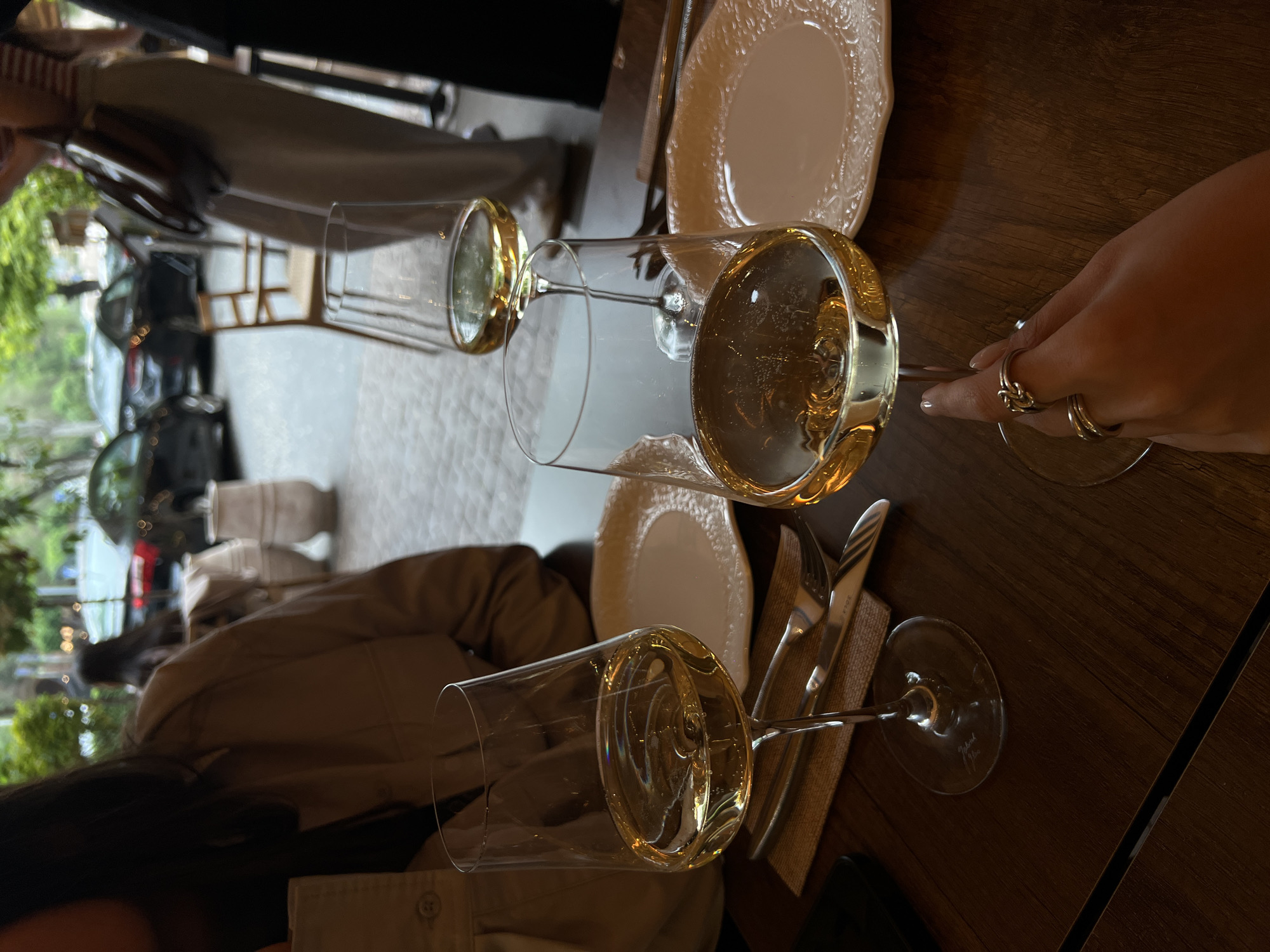 The image captures a dining scene in Yerevan, Armenia. Two wine glasses filled with white wine sit on a dark wooden table, accompanied by plates, silverware, and linen napkins. A person's hand, adorned with rings, holds one of the glasses. Outside the window, glimpses of the city are visible, featuring trees, buildings, and parked cars. The overall setting suggests a restaurant or cafe setting.