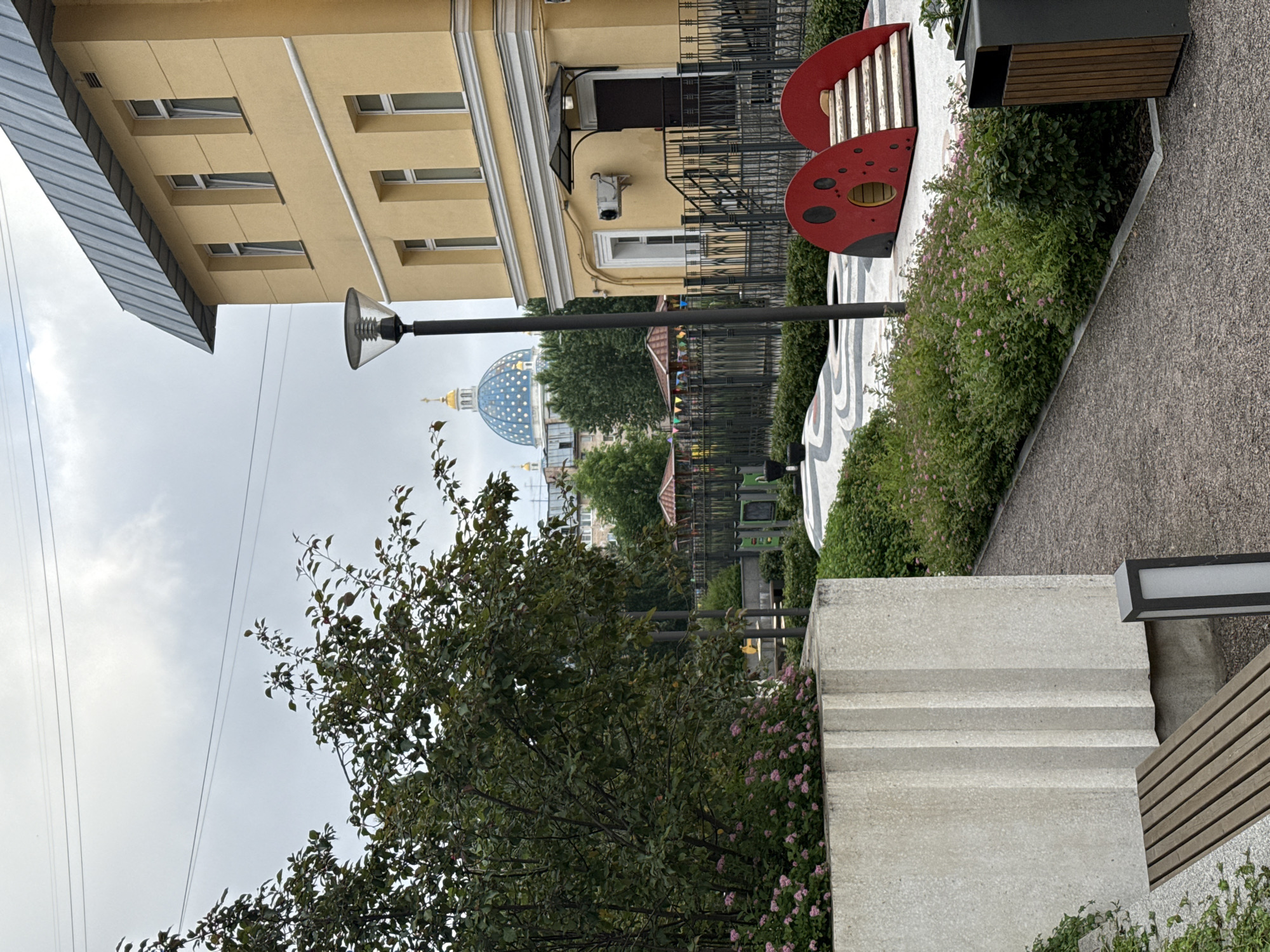 The image depicts a courtyard area in Saint Petersburg, Russia, featuring a playground and a yellow building with a grey metal roof. In the background, a prominent blue dome with golden accents, characteristic of a church or cathedral, is visible. The foreground includes a concrete planter with lush green foliage and pink flowers, a wooden bench, and a gravel pathway. A distinctive red ladybug-shaped playground structure with steps is also present. A tall street lamp stands near the building. The sky is overcast.