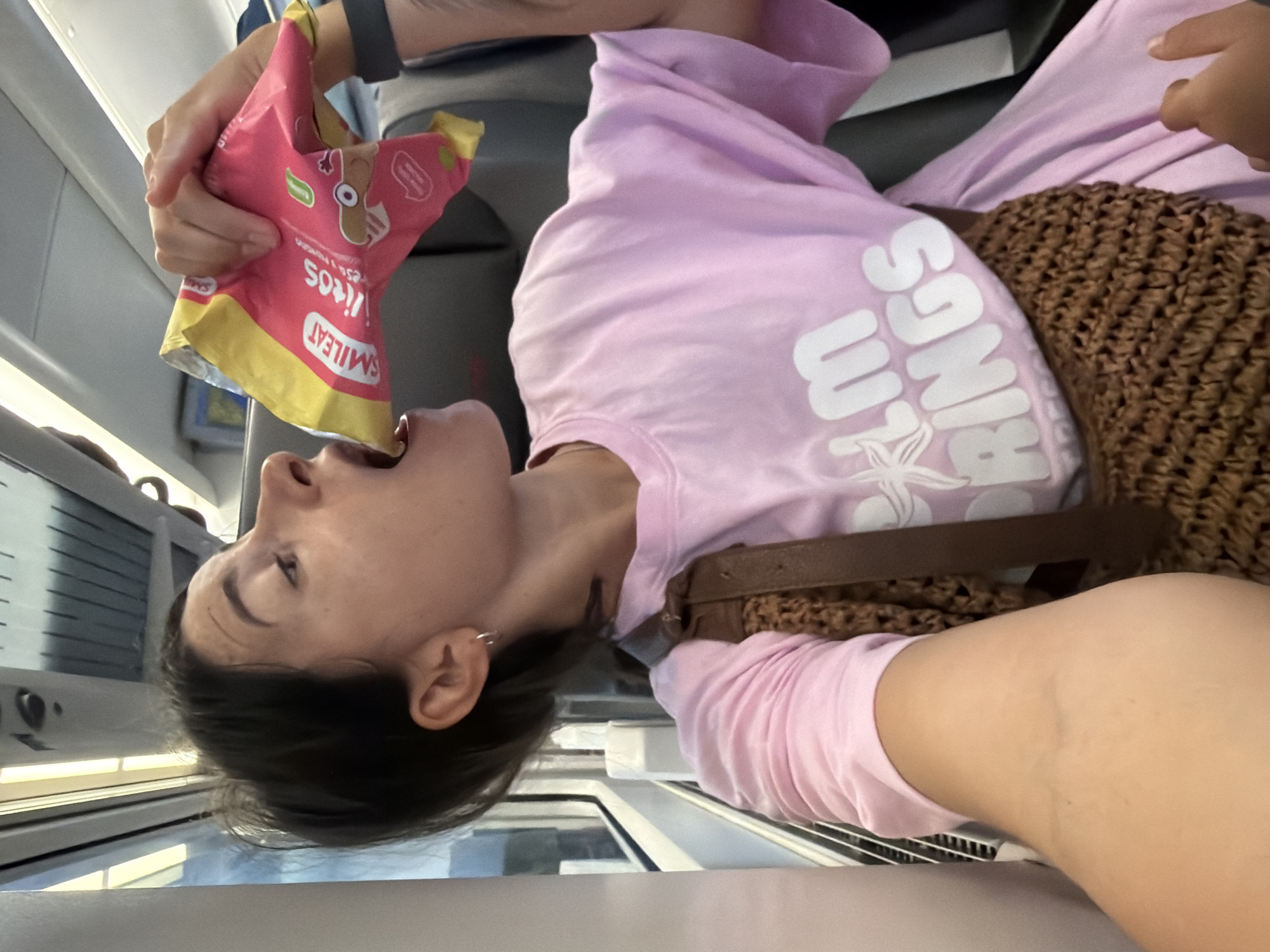 The image depicts a woman inside a train carriage, in the city of Tarragona, Spain. She is wearing a pink t-shirt with the text "PALM SPRINGS" printed in white. A woven brown bag is slung over her shoulder, resting on her chest and stomach. The woman is in the process of eating from a pink and yellow bag of snacks, holding it up to her mouth. The interior of the train carriage is visible, with seats and windows. The lighting suggests it is daytime.