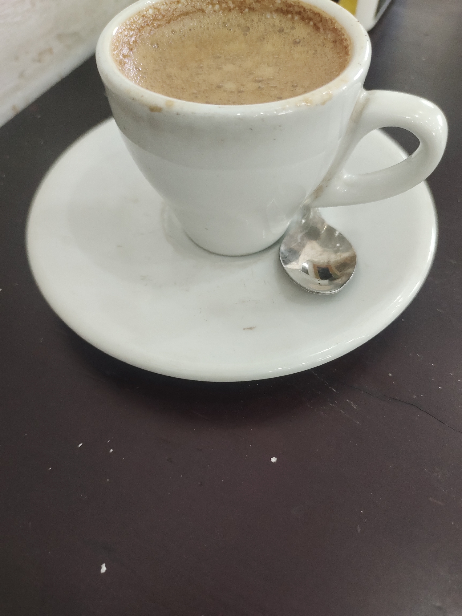 The image depicts a white porcelain cup and saucer containing a coffee beverage, likely espresso or a similar style, with a thin layer of crema visible. A small silver spoon rests on the saucer next to the cup. The cup and saucer are placed on a dark brown table or countertop. The background is out of focus. The photo was taken in Lhoksukon, Indonesia.