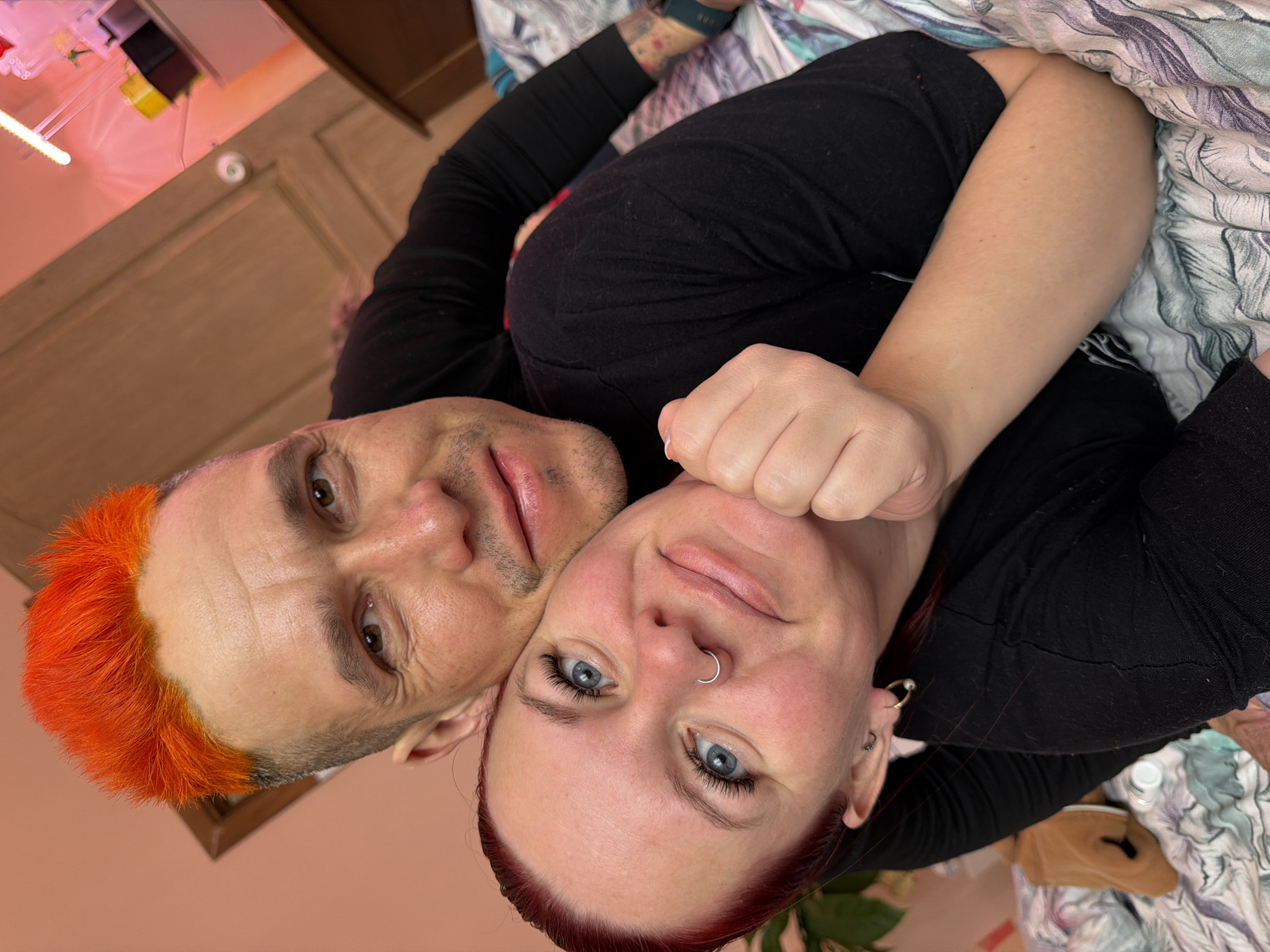 The image shows a man and a woman lying together, possibly on a bed. The man has short, orange hair, while the woman has red hair and a nose ring. Both are wearing black tops. The background includes a patterned bedspread and furniture. The location is unknown; there is no indication the location is Chaikovsky, Russia.