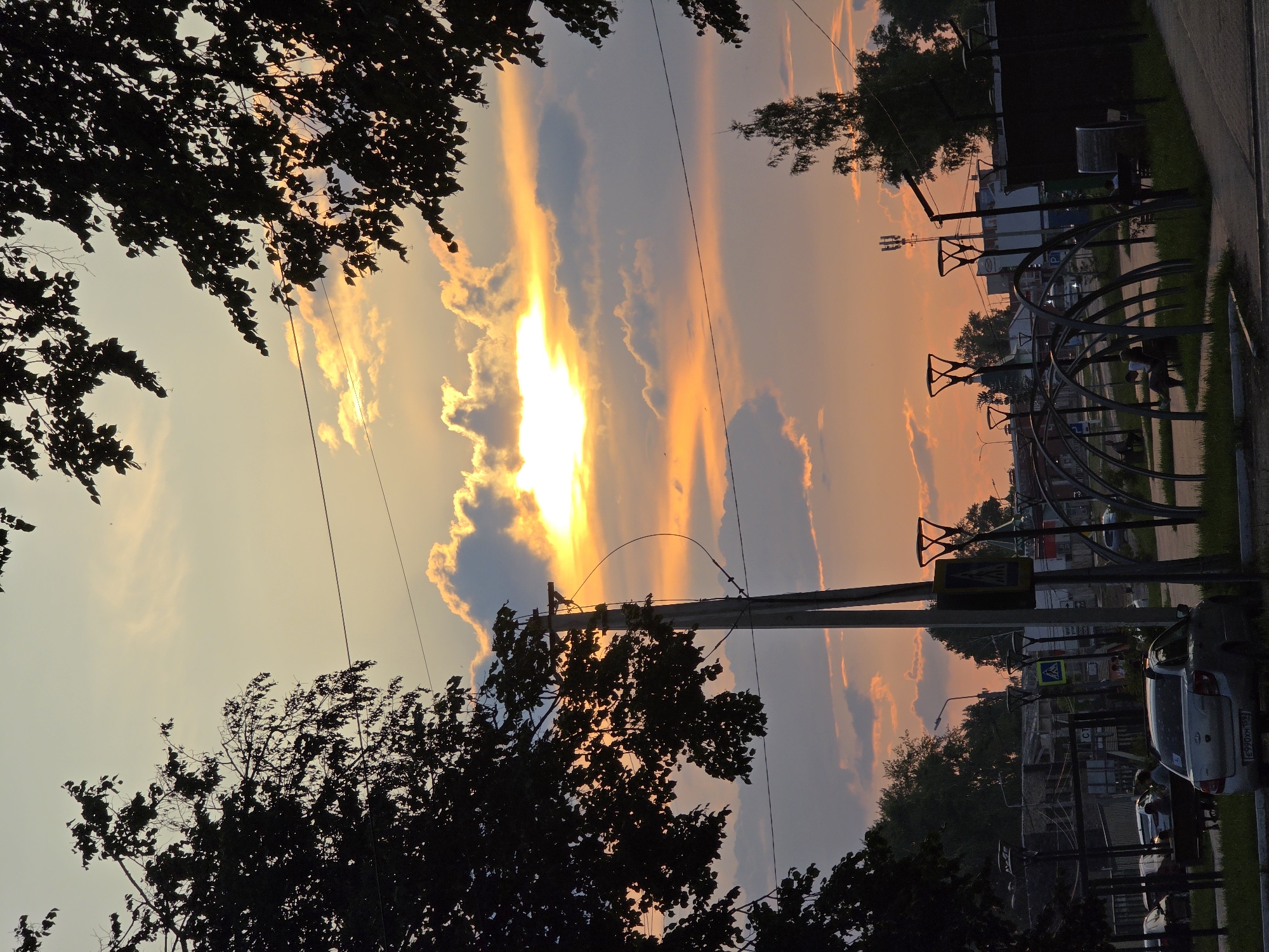 The image captures a sunset scene in Chaikovsky, Russia. The sky is filled with warm hues of orange and yellow as the sun sets behind clouds. Silhouetted trees frame the top of the image. In the foreground, there is a street with a silver sedan parked on the right. A utility pole with wires stands prominently in the mid-ground. Several people are visible sitting on benches in a park-like area with arched structures. Buildings and other urban elements are present in the background, blending into the twilight atmosphere.