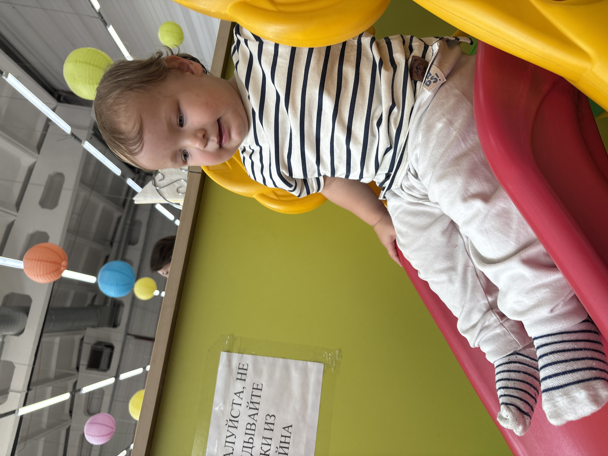 In the city of Chaikovsky, Russia, a young child with blond hair and blue eyes is seated on a red and yellow plastic slide indoors. The child is wearing a white t-shirt with navy blue horizontal stripes and light-colored pants, along with white socks with navy blue stripes. The child is looking to the right with a slight smile. Behind the child, a bright green wall is visible with a white sign containing Russian text. Several colorful paper lanterns, including orange, pink, and blue, are suspended from the ceiling above. The environment appears to be a children's play area or indoor playground.