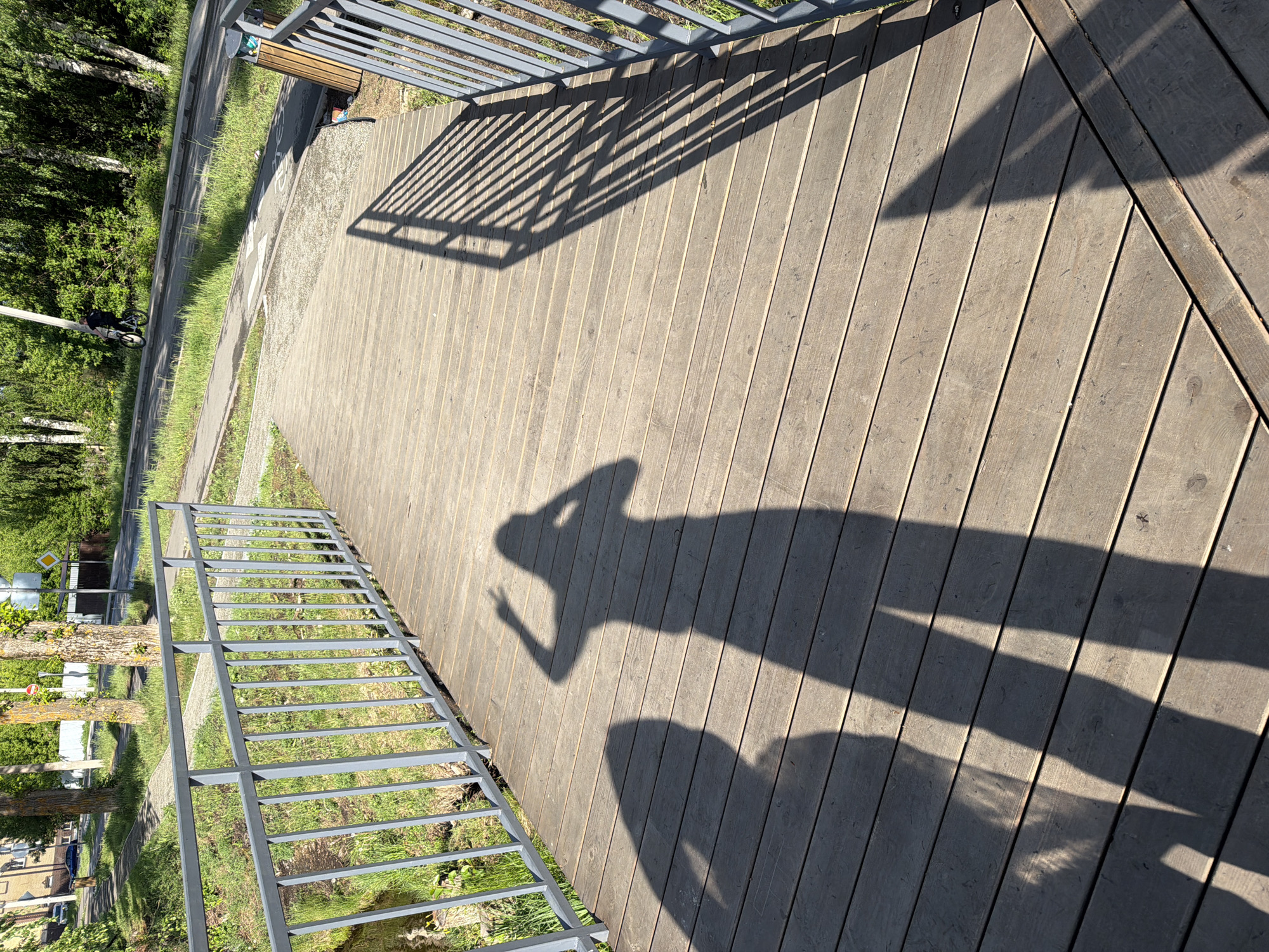 The image shows a wooden deck in Votkinsk, Russia. The shadow of a person taking a photo is cast upon the deck. A metal railing borders the deck on two sides. A street and trees are visible beyond the deck and railing. In the background, a cyclist can be seen on the pathway.