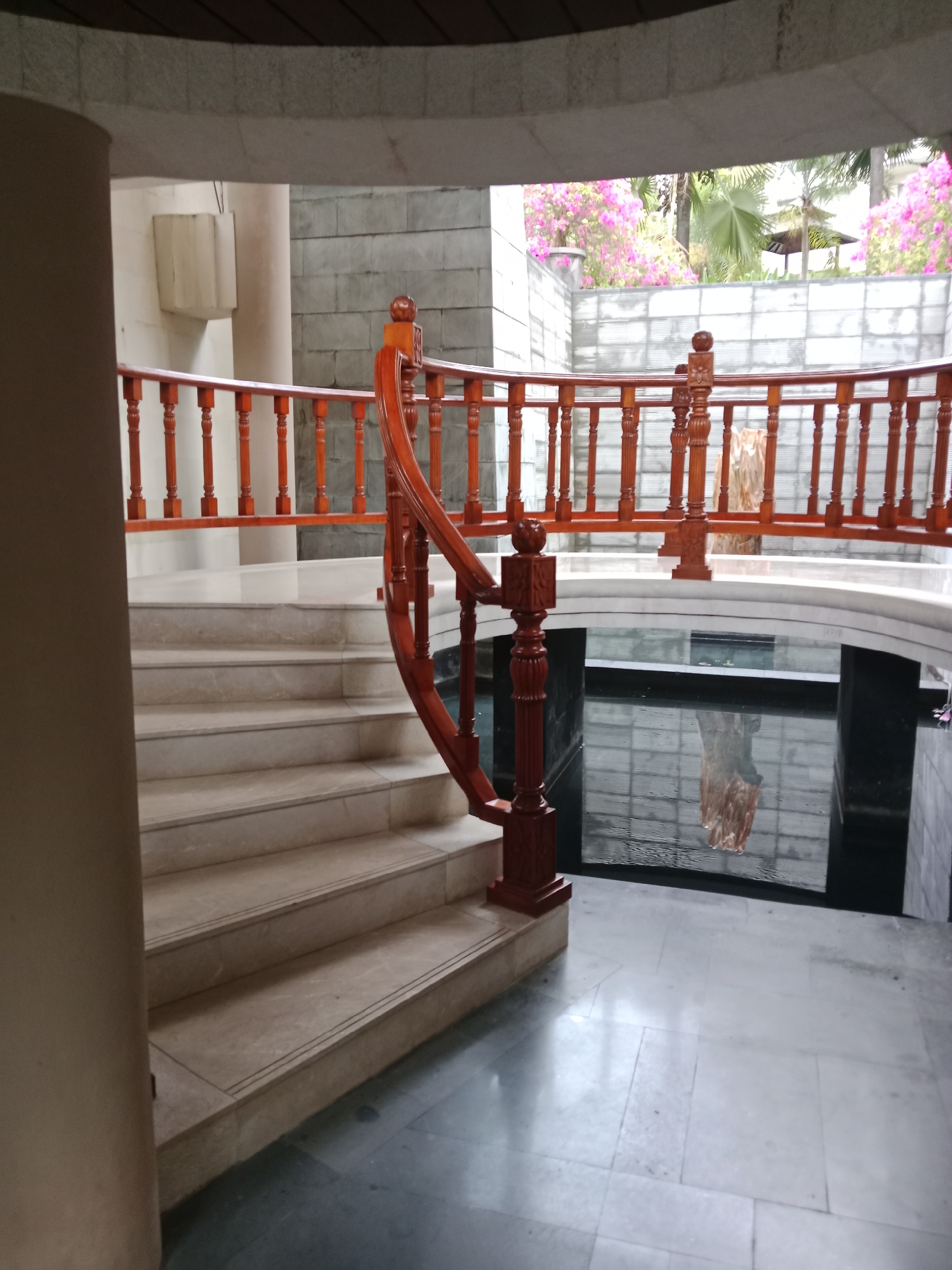 The image shows an interior shot of a building with a curving wooden staircase and railing. The staircase is made of light-colored marble. Below the staircase, there is a dark pool of water with a reflection of what appears to be a statue or decorative object. In the background, there is an outdoor area with pink flowers and palm trees visible through a glass or open wall. The overall architecture suggests a resort or hotel lobby in Kuta Selatan, Indonesia.