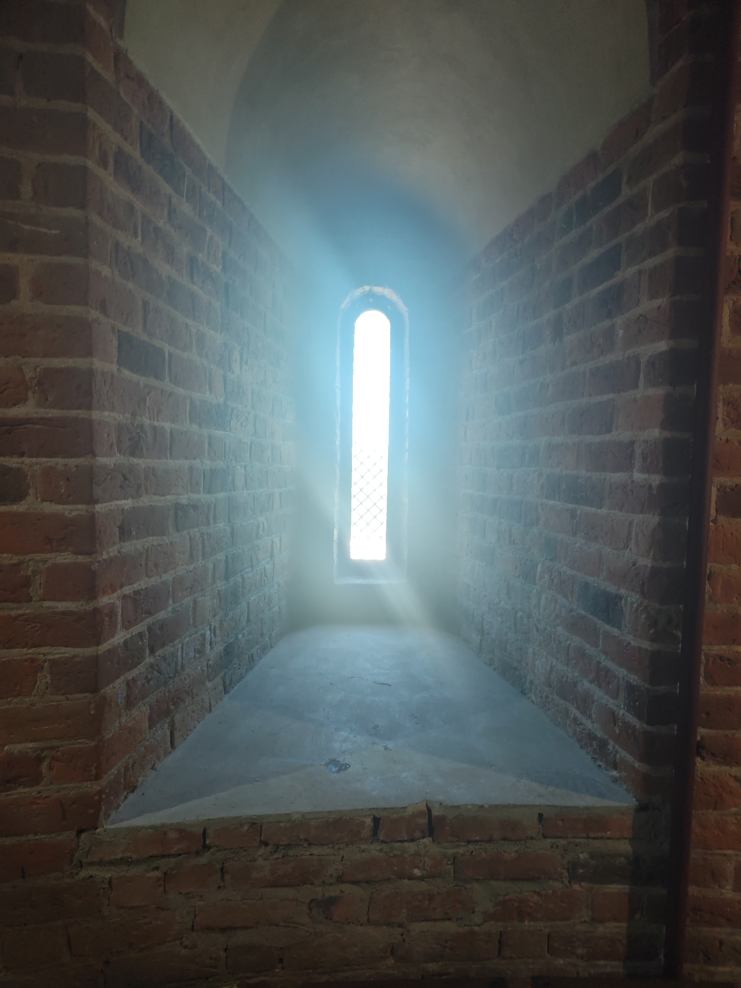 A narrow, arched window pierces a thick brick wall, allowing a bright shaft of light to stream into a recessed space. The light illuminates dust particles in the air, creating a hazy glow that softly lights the grey concrete floor of the recess. The walls of the recess are also made of brick, with the same warm, reddish-brown hue as the outer wall. The scene is likely part of a historical building, possibly a fortification or an old church, given the sturdy construction and the architectural style. The location is the city of Kamyanyets, Belarus, where such structures are common.