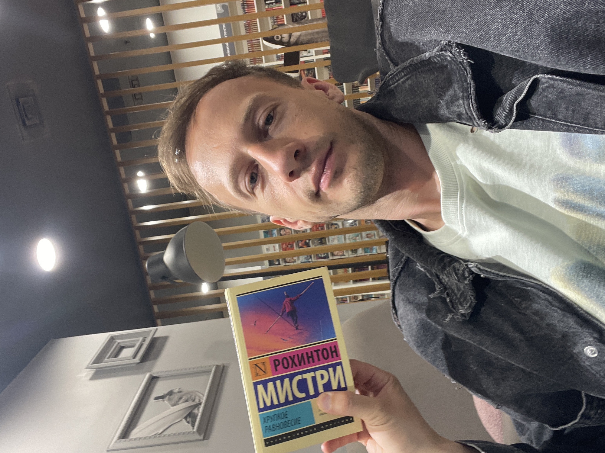 A man is holding a book titled "Мистри" by Р. Рохинтон. He is wearing a dark denim jacket and a light-colored t-shirt. The background appears to be a bookstore, with shelves of books visible behind him. The lighting suggests it is indoors, possibly in the evening. The man is looking at the camera, taking a selfie. There are framed pictures on the wall to his left and a modern-style lamp hanging from the ceiling. The overall setting is casual and relaxed.