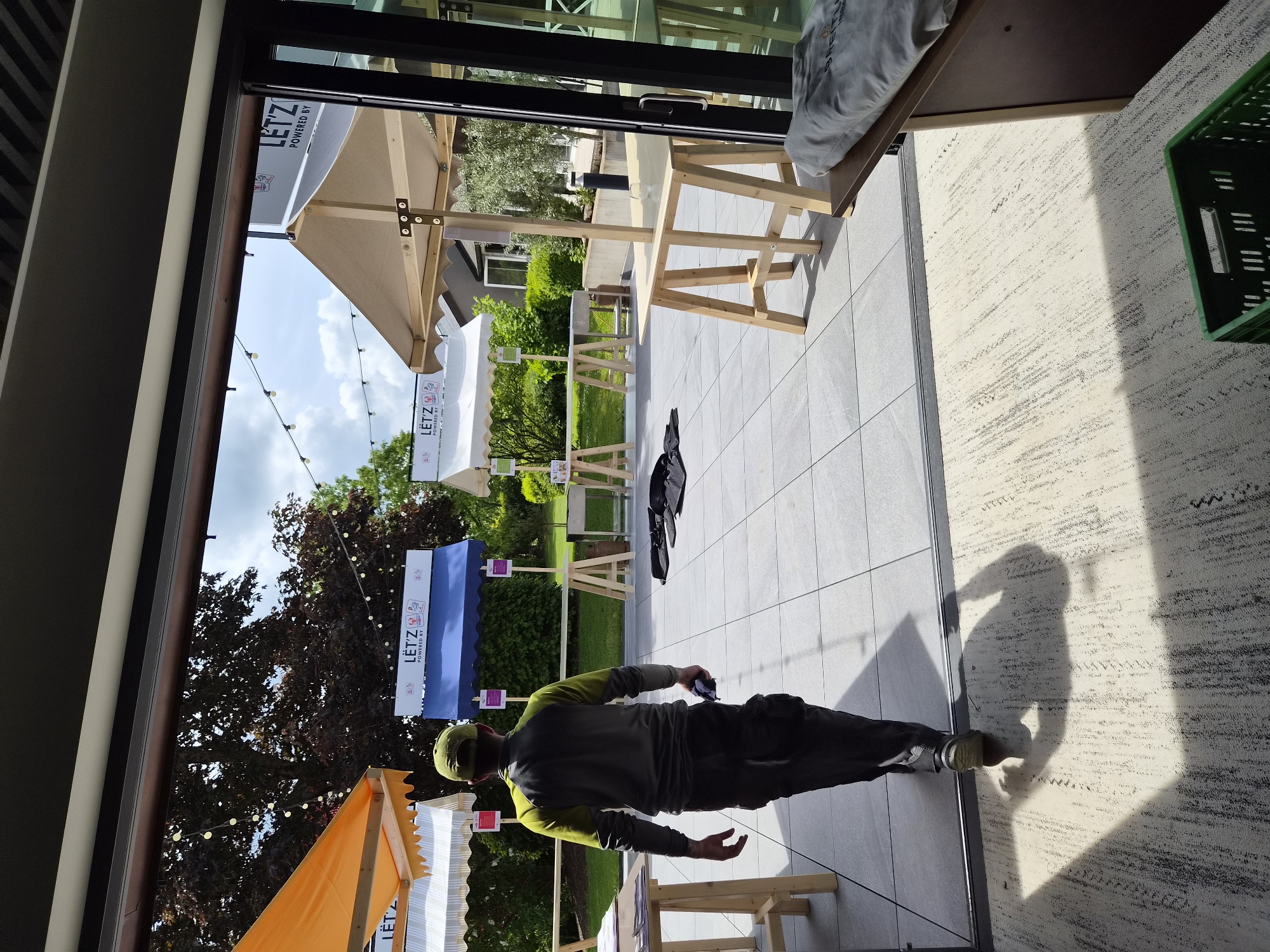 A man wearing a grey long-sleeved shirt with neon green sleeves, dark trousers, and a green baseball cap walks on grey tiles outdoors. He is holding a small dark object in his right hand. The setting appears to be an event preparation area in Lenningen, Luxembourg, evidenced by the presence of tents labeled "LET'Z POWERED BY" and wooden table frames. Trees and landscaping define the background. The photo is taken from inside a building, with a partial view of the interior visible. A shadow is cast on the carpeted floor inside the building.