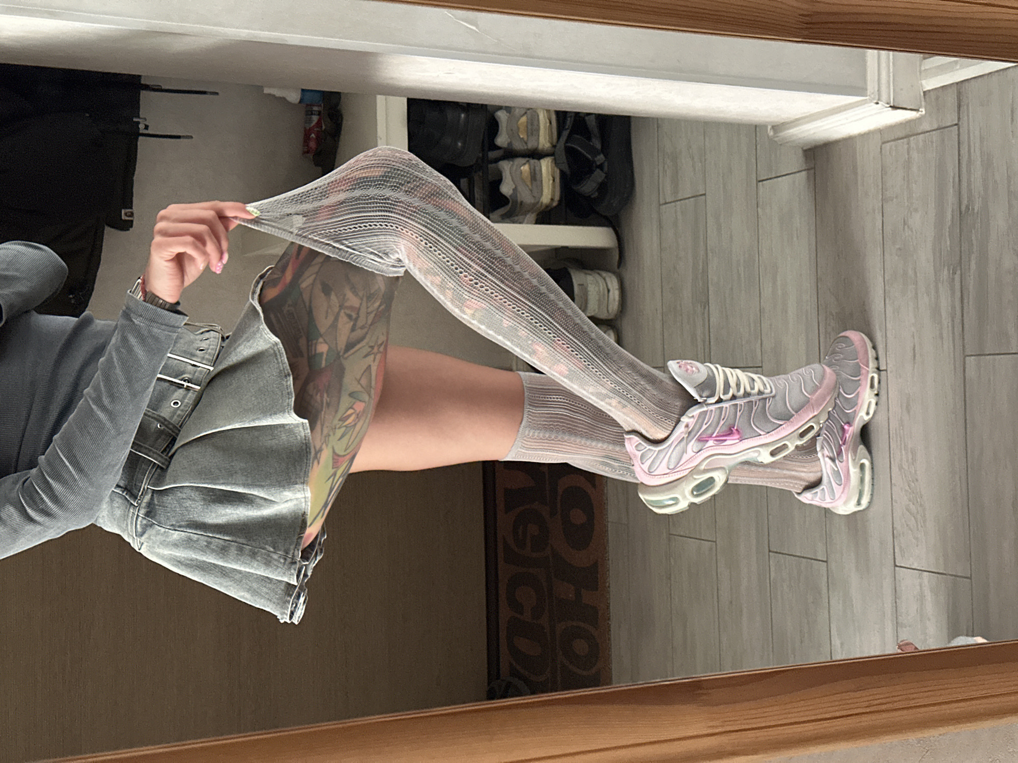 The media file is a vertically oriented, full-body reflection of a person in a mirror. The person is wearing a gray, long-sleeved top, a gray denim skirt with a belt, patterned gray and pink thigh-high socks, and gray and pink athletic shoes. The person's left arm is bent, and their hand is pulling at the top of the thigh-high sock, showing the texture and stretch of the garment. The person has visible tattoos on their left leg, which are partially obscured by the sock and skirt.
The background appears to be an indoor setting, possibly a closet or dressing area, with shelves containing shoes visible in the reflection. The floor is tiled in a light gray wood-grain pattern. The reflection is framed by a wooden border, suggesting the mirror is set within furniture or a doorway.
The lighting is soft and appears to be artificial, consistent with an indoor environment. There are no strong shadows, indicating an even light source. The overall mood is casual and possibly indicative of getting dressed or posing for a photo.
No discernible time of day or weather conditions are evident. The location context is Moscow, Russia, but there are no specific visual cues to confirm this within the image itself.
Visible text is limited to the pattern of the socks and a dark mat with large, light-colored lettering, which reads "O HO" and possibly more text below, reflected on the floor.