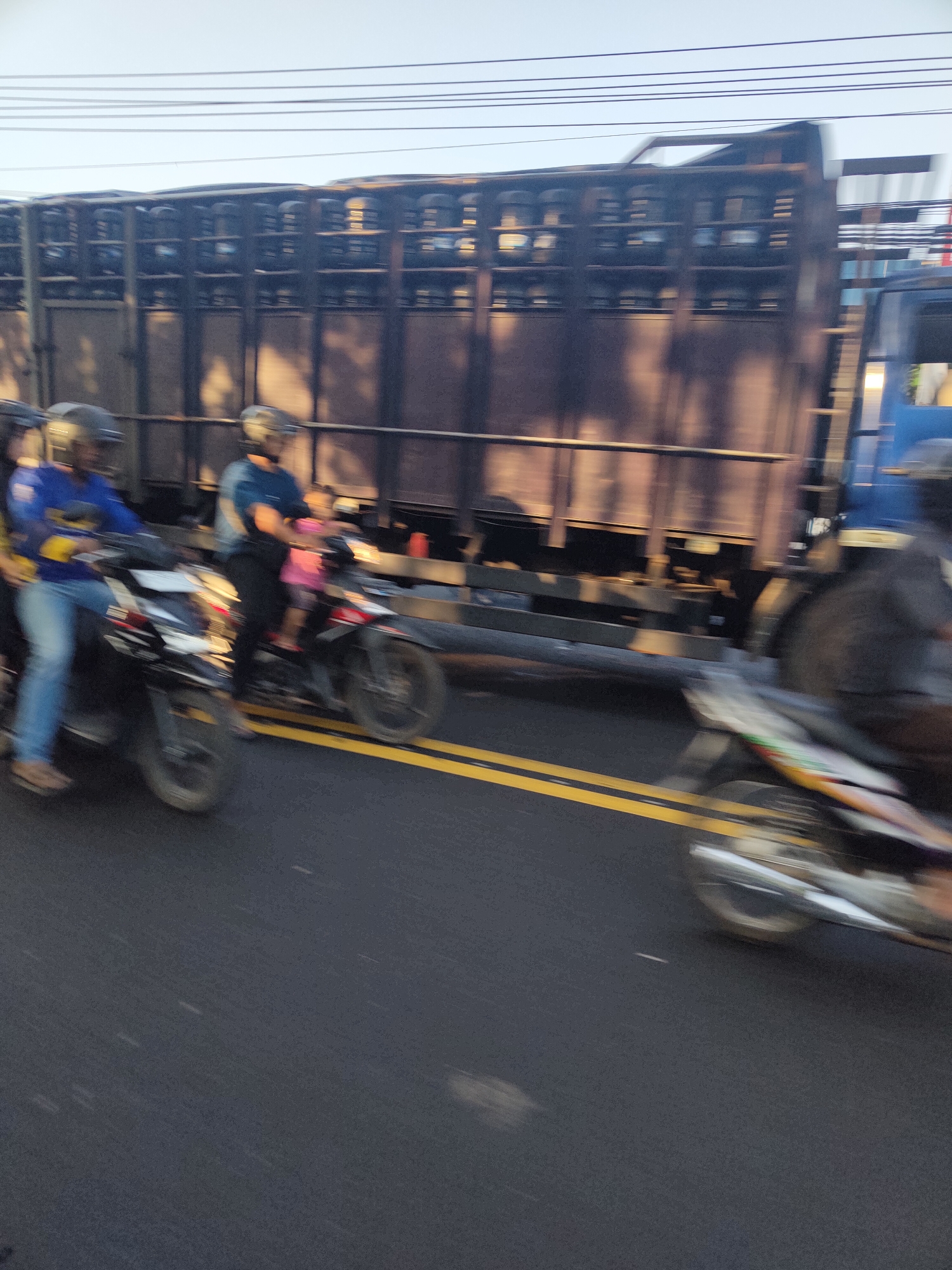 The image shows a busy street scene in Dermo, Indonesia, with several motorcycles in motion. In the foreground, two motorcycles are visible, one with two riders, a driver and a passenger, and another motorcycle with a single rider. The motorcycles are on a paved road marked with yellow double lines. Behind the motorcycles, a large truck with a dark, corrugated metal cargo area is prominent, obscuring much of the background. Power lines are visible against the sky. The lighting suggests it is either early morning or late afternoon. The motion blur indicates that the photograph was taken while the vehicles were moving.