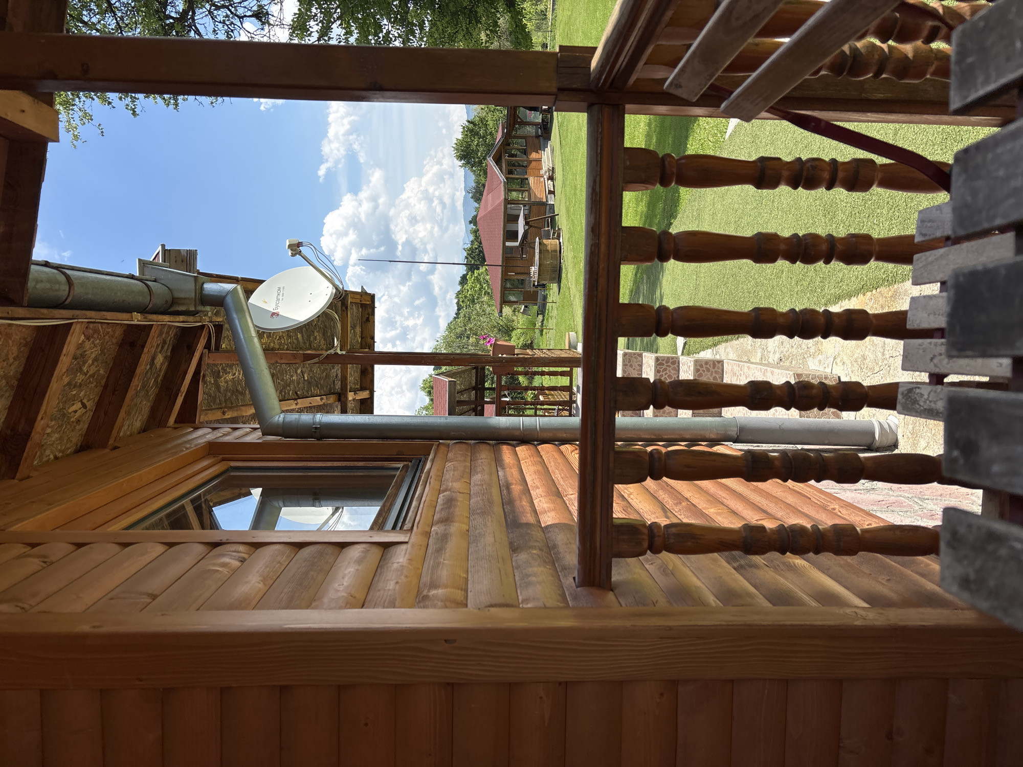 The image depicts a static outdoor scene captured from the perspective of a wooden porch or balcony, likely at a cabin or lodge in Zasele, Bulgaria. The immediate foreground shows the interior of this wooden structure, with a partial view of a rustic wooden table or bench in the bottom-center. A prominent wooden balustrade and handrail, crafted with turned balusters, extends across the lower portion of the frame, defining the edge of the porch.
To the left, the exterior wall of the building is constructed from horizontal logs, showcasing a light brown wood grain. A window with dark framing is set into this wall. Above the window, a gray metal rain gutter system is visible, running horizontally and then descending vertically. Attached below the gutter, a white satellite dish, bearing the "Бусатком" (Bulsatcom) logo, is mounted and points upwards towards the sky. The overhang of the porch roof is visible at the top, constructed with exposed wooden beams and plywood sheathing.
Beyond the porch and railing, a vibrant green lawn, possibly artificial turf or meticulously maintained grass, stretches into the midground. In the center of the midground, a substantial single-story wooden building with a prominent red tile roof stands. This building features numerous windows and a covered porch area. To its left, a circular wooden structure, resembling a hot tub or a large planter, is situated on the lawn, adjacent to a wooden swing set. A tall, thin metal pole is visible behind the hot tub structure, extending upwards into the sky.
The background consists of rolling hills or mountains covered with dense green foliage, indicating a rural or semi-forested environment. The sky above is clear blue with scattered, fluffy white cumulus clouds. No people or dynamic actions are present in the frame.