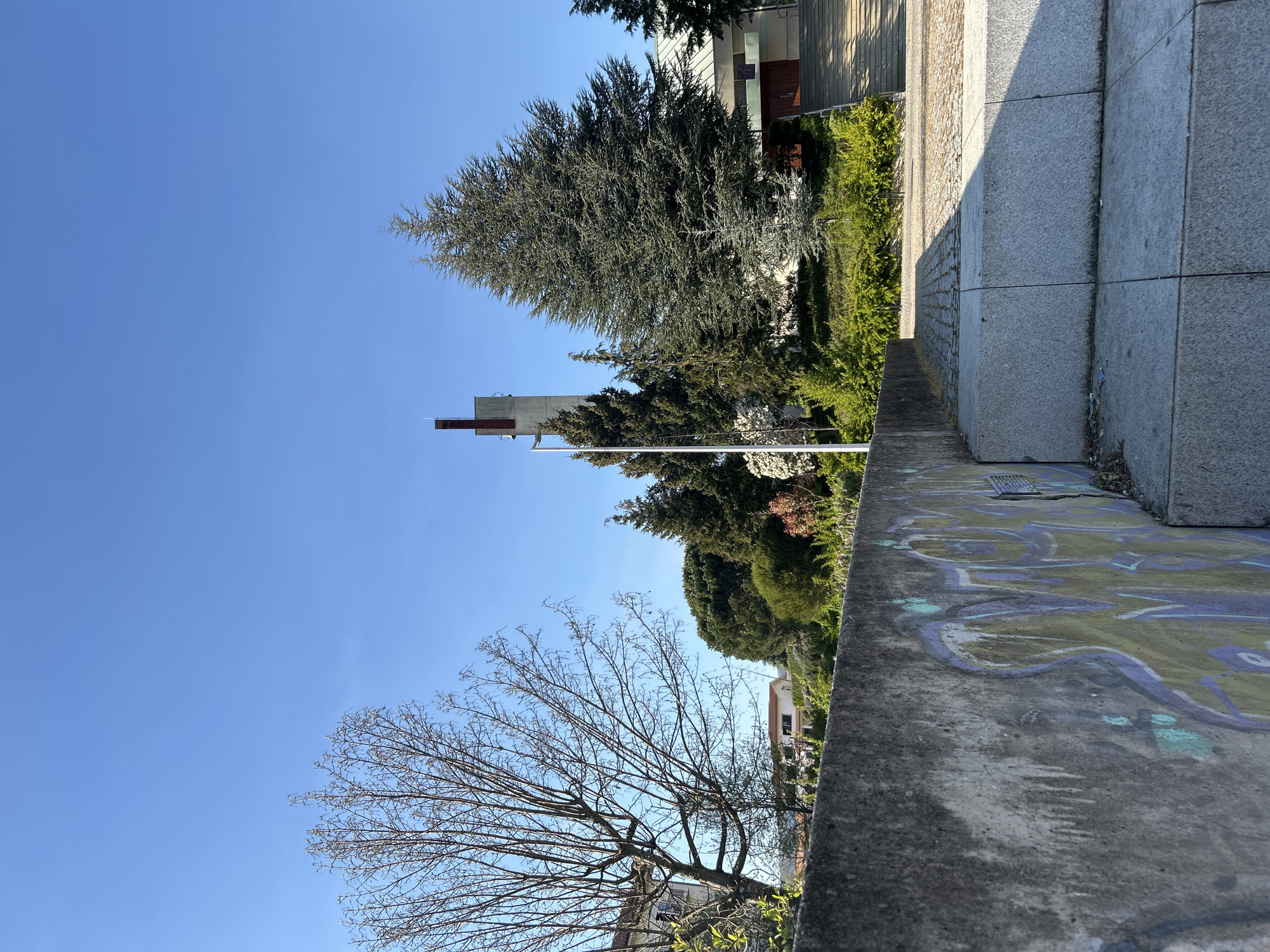 The image shows an outdoor scene in Vila Real, Portugal, featuring a concrete wall with graffiti. In the background, there are trees and a church building, with a clear blue sky above. A bare tree is visible on the left side of the image. The photograph is taken from a low angle, emphasizing the concrete wall and the objects in the background. The overall environment appears to be urban.